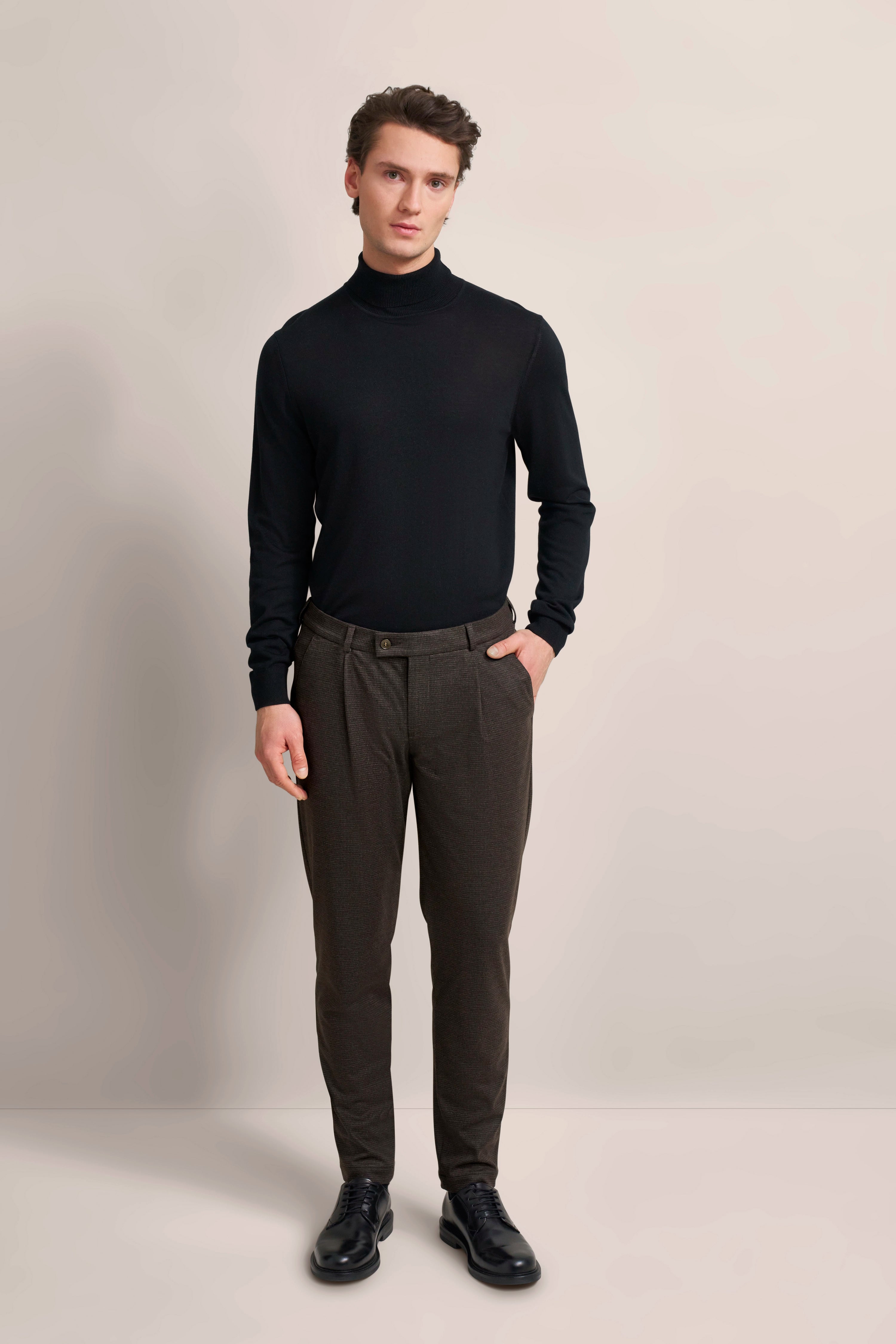 Model in schwarzem Rollkragenpullover, brauner Hose, schwarzer Schuhen. Stehende Pose. bugatti Stil.