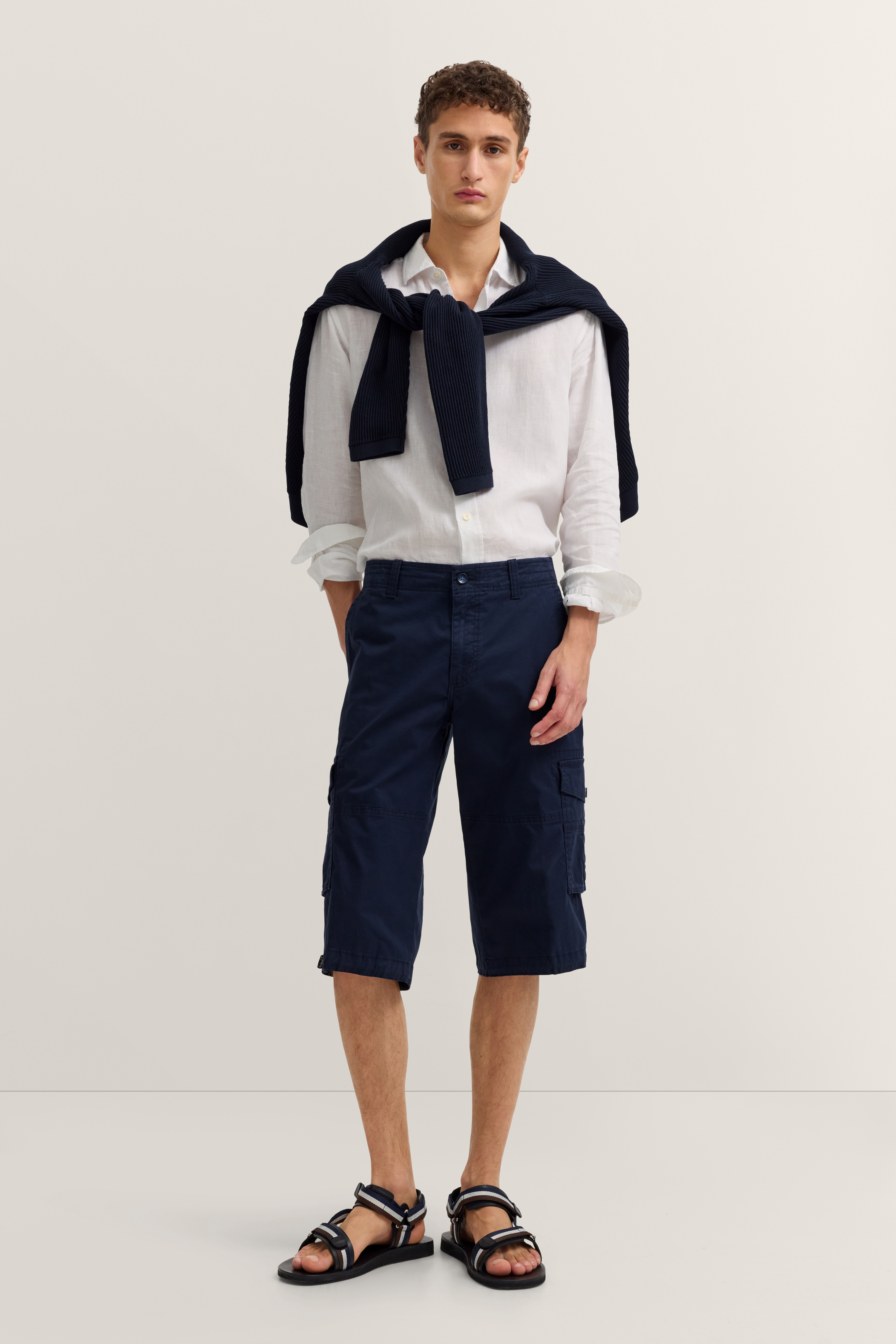 Model in weißem Hemd, dunkelblauer Shorts und Pullover über Schultern, lässige Pose, bugatti Stil.