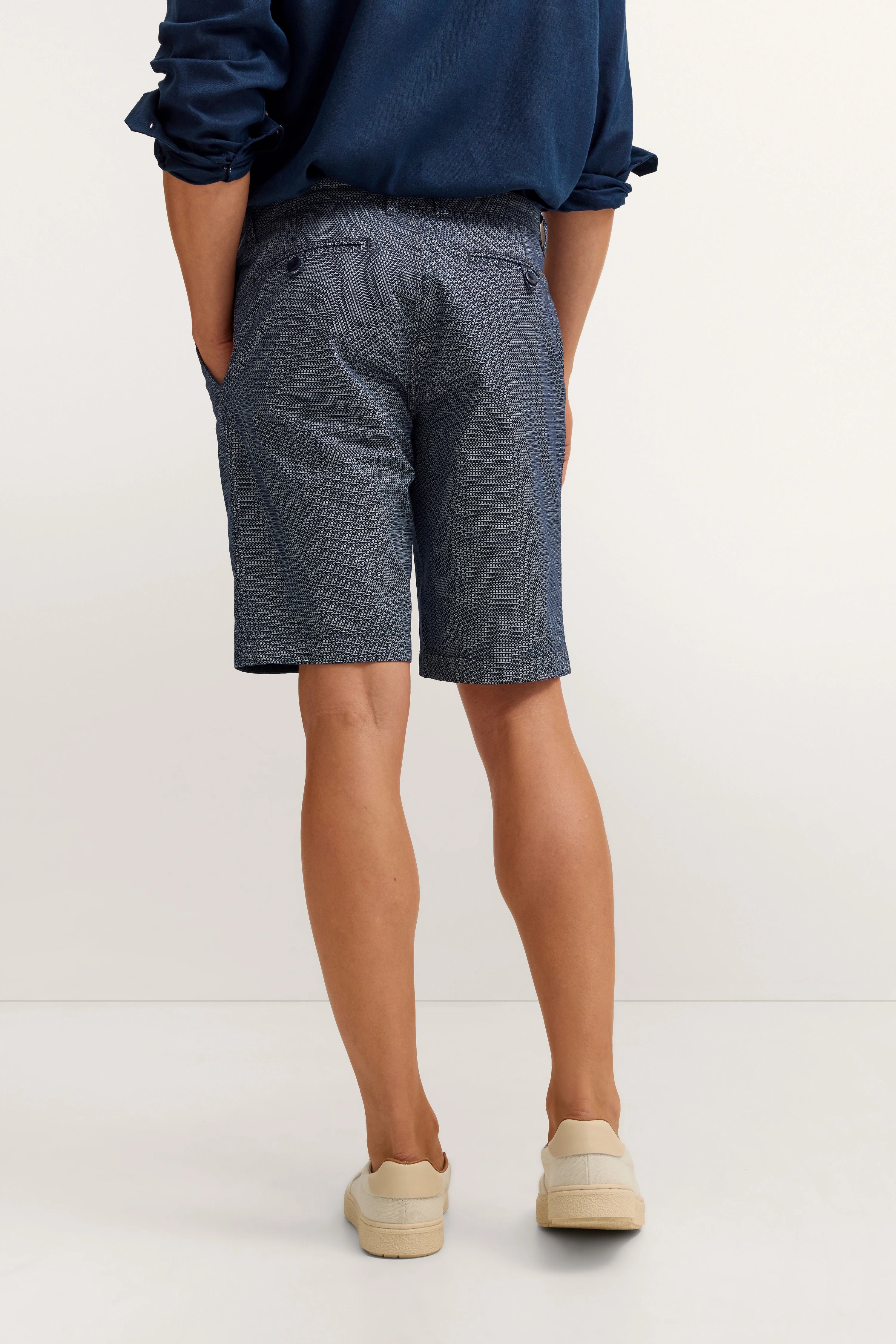 Bugatti Modelbild, blaues Hemd, graue gemusterte Shorts, beige Sneaker, entspanntes Posing von hinten.