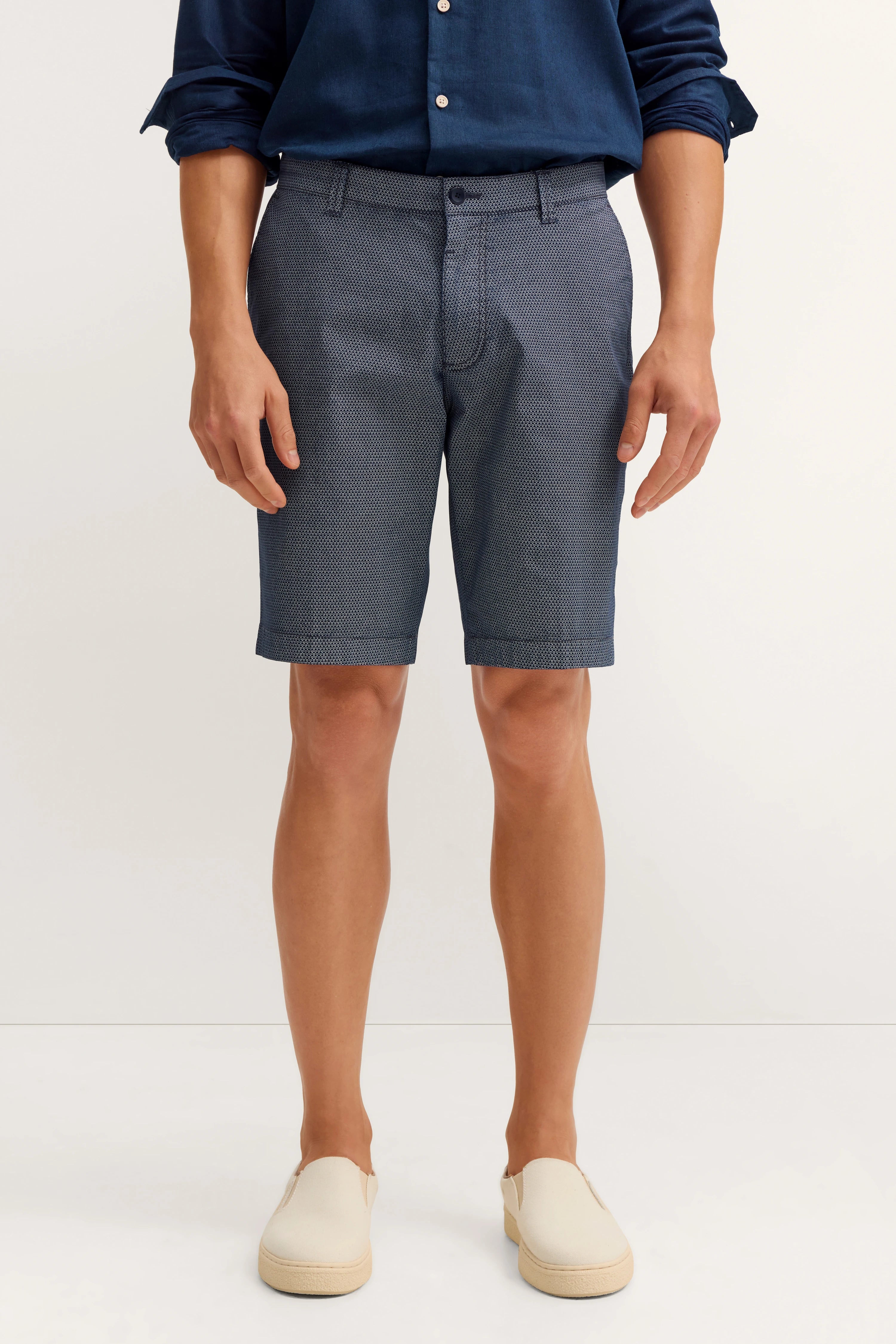 Bugatti Modelbild, relaxed Pose, dunkelblaues Hemd, graue gemusterte Shorts, cremefarbene Slip-Ons.
