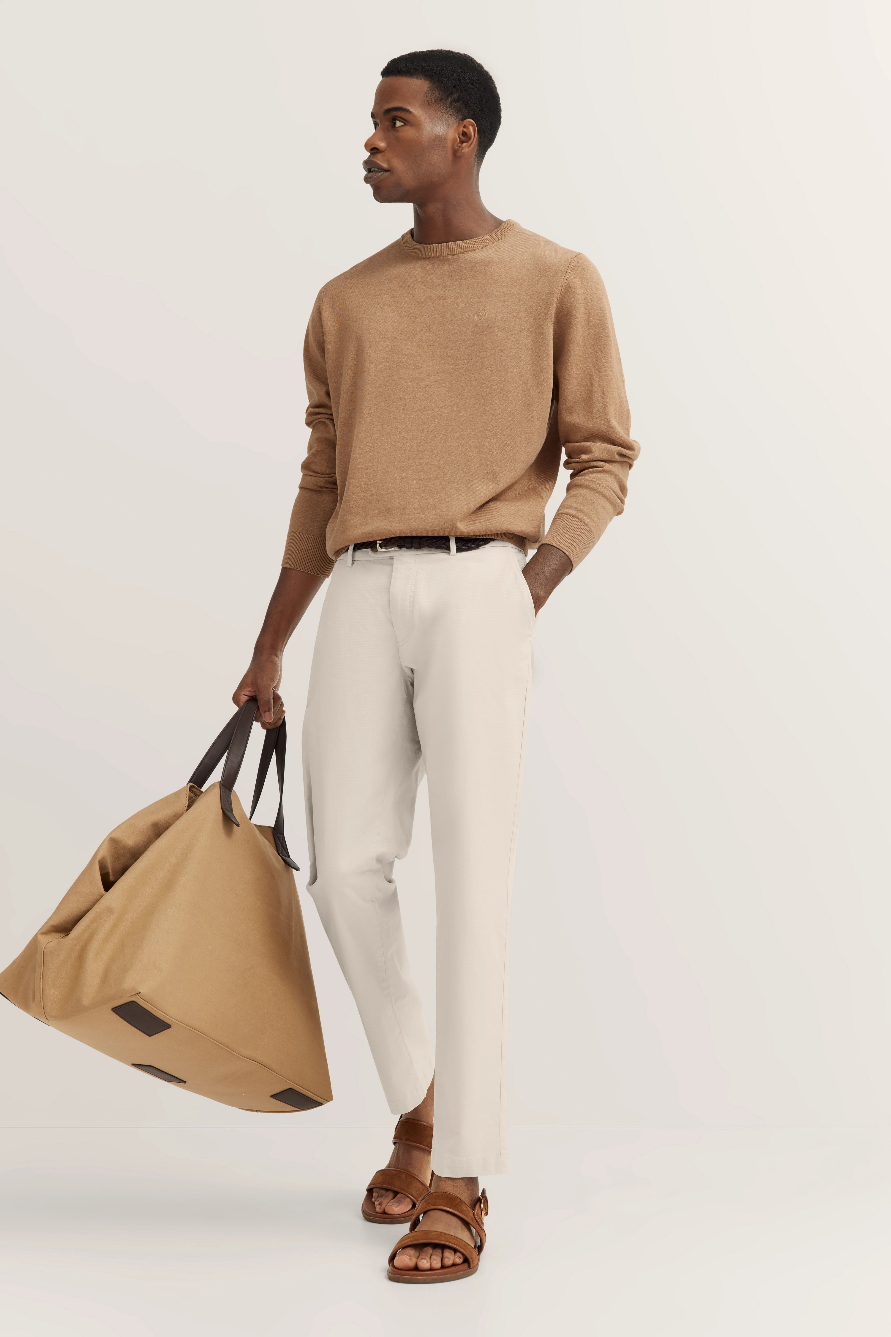Beige trui, witte broek, bruine sandalen, model met tas in ontspannen pose.