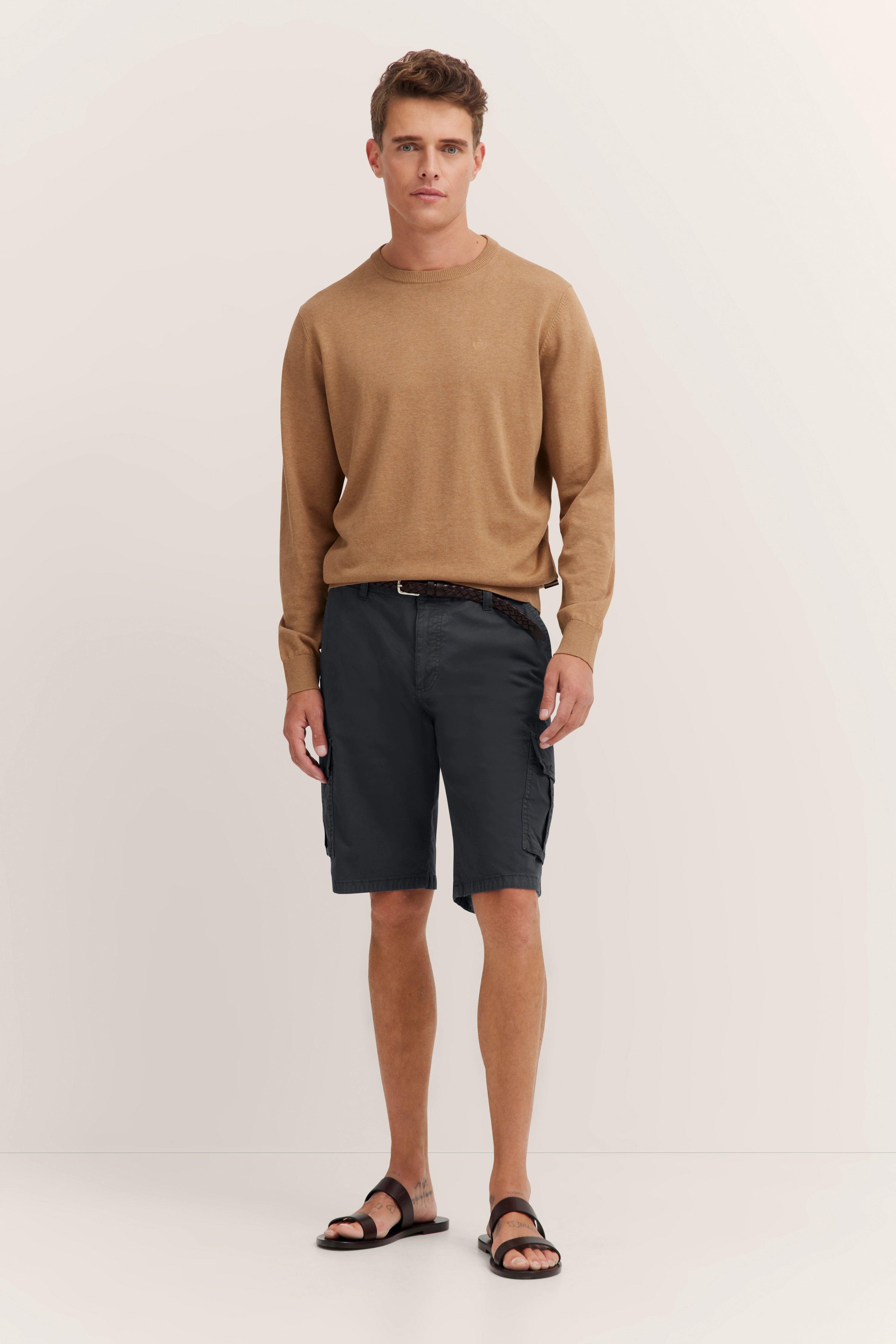 Modelbild mit braunem Pullover, grauen Shorts und Sandalen, entspannte Pose. bugatti Stil.