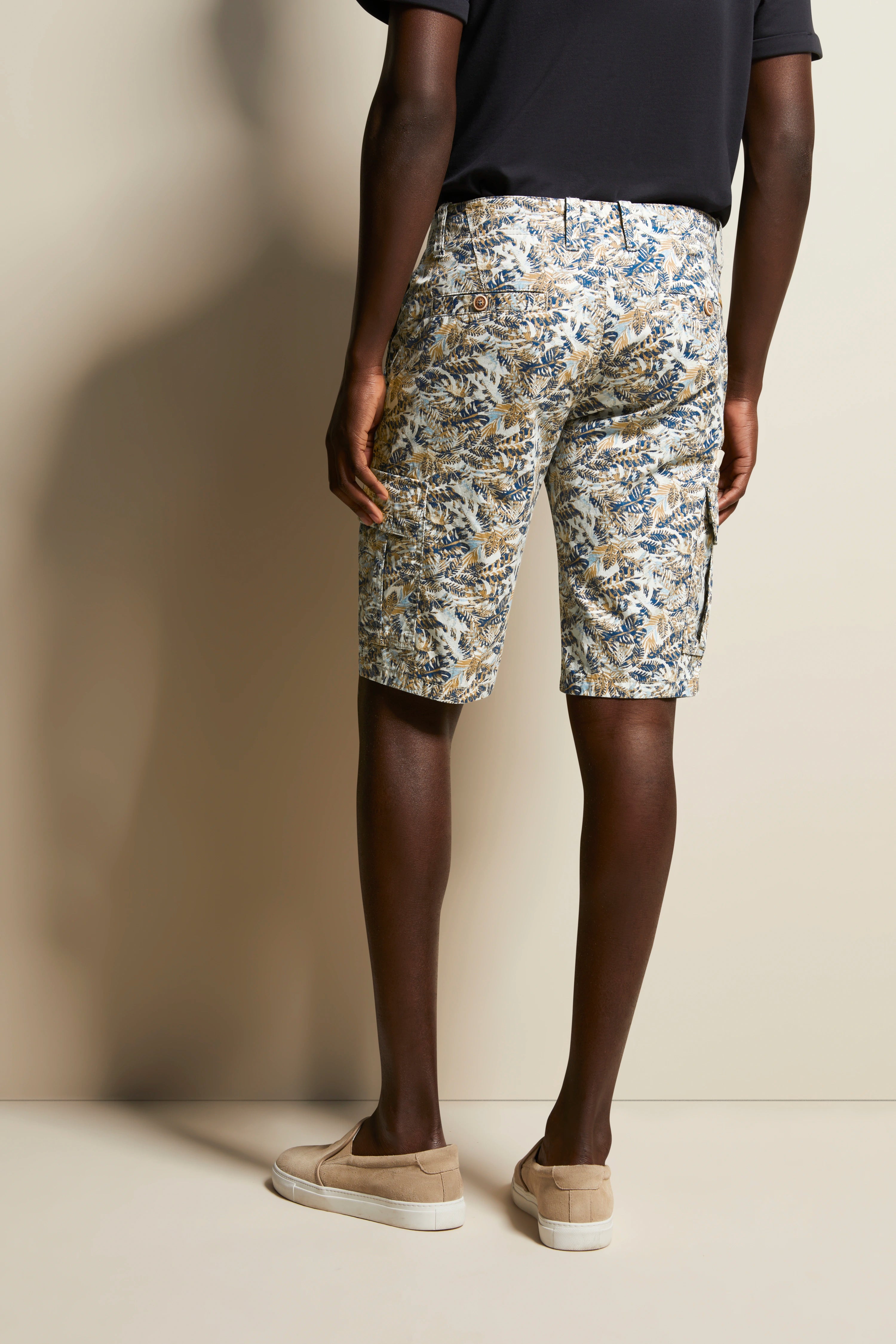 bugatti Modelbild, florale gemusterte Shorts in Beige-Blau, lässige Pose von hinten, beige Schuhe.