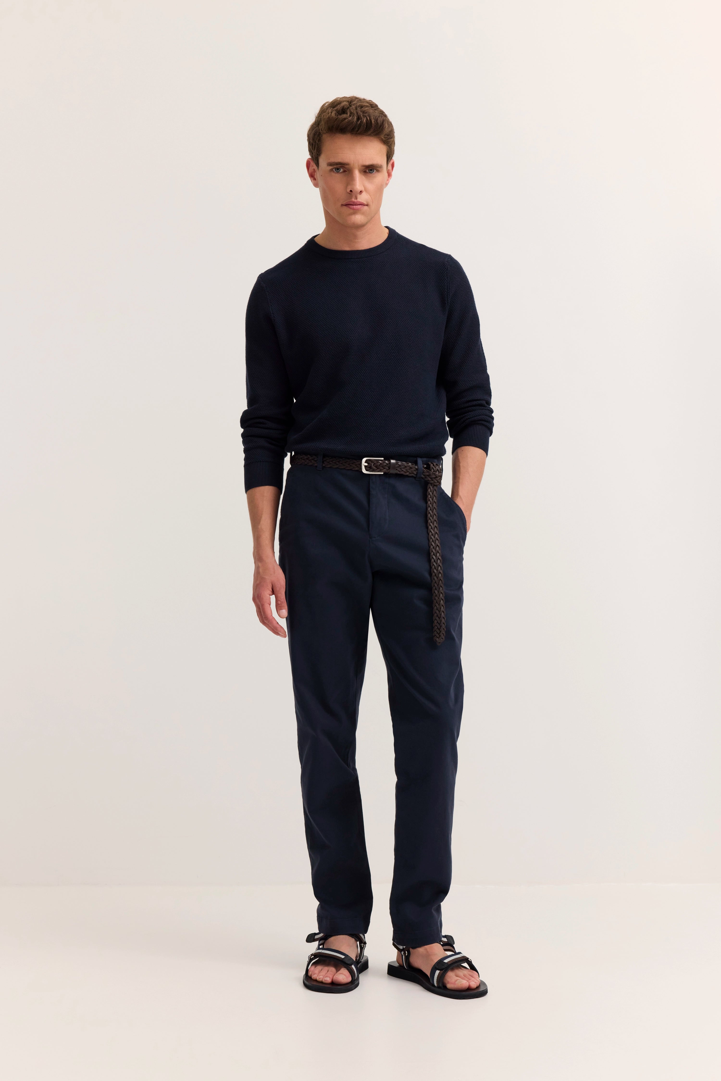 Bugatti Modelbild: Dunkler Pullover, dunkelblaue Hose, schwarze Sandalen. Entspanntes Posing mit Hand in Tasche.