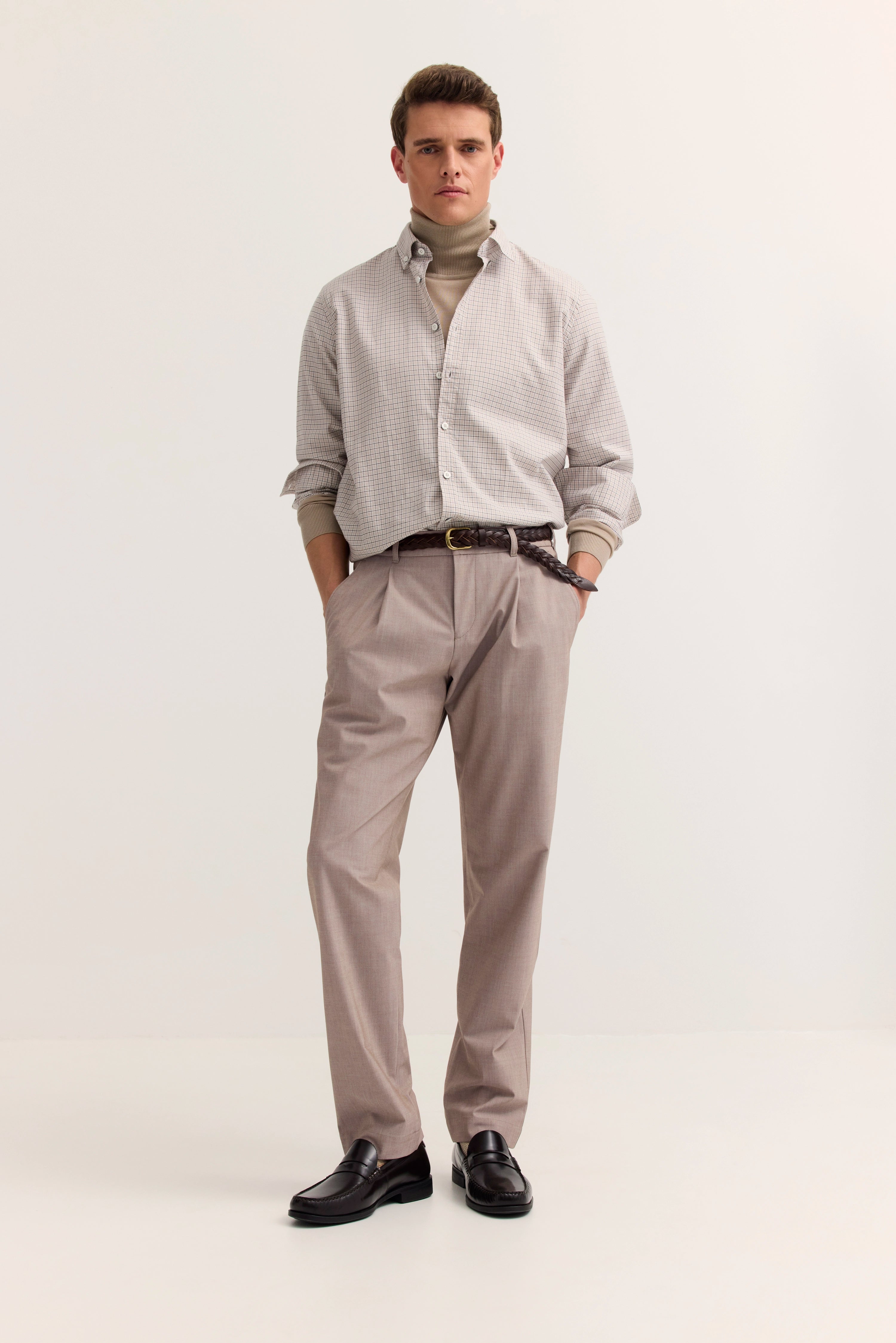 Model in beige gemusterter bugatti-Hemd, Rollkragenpullover, helle Hose, brauner Gürtel, entspannte Pose.