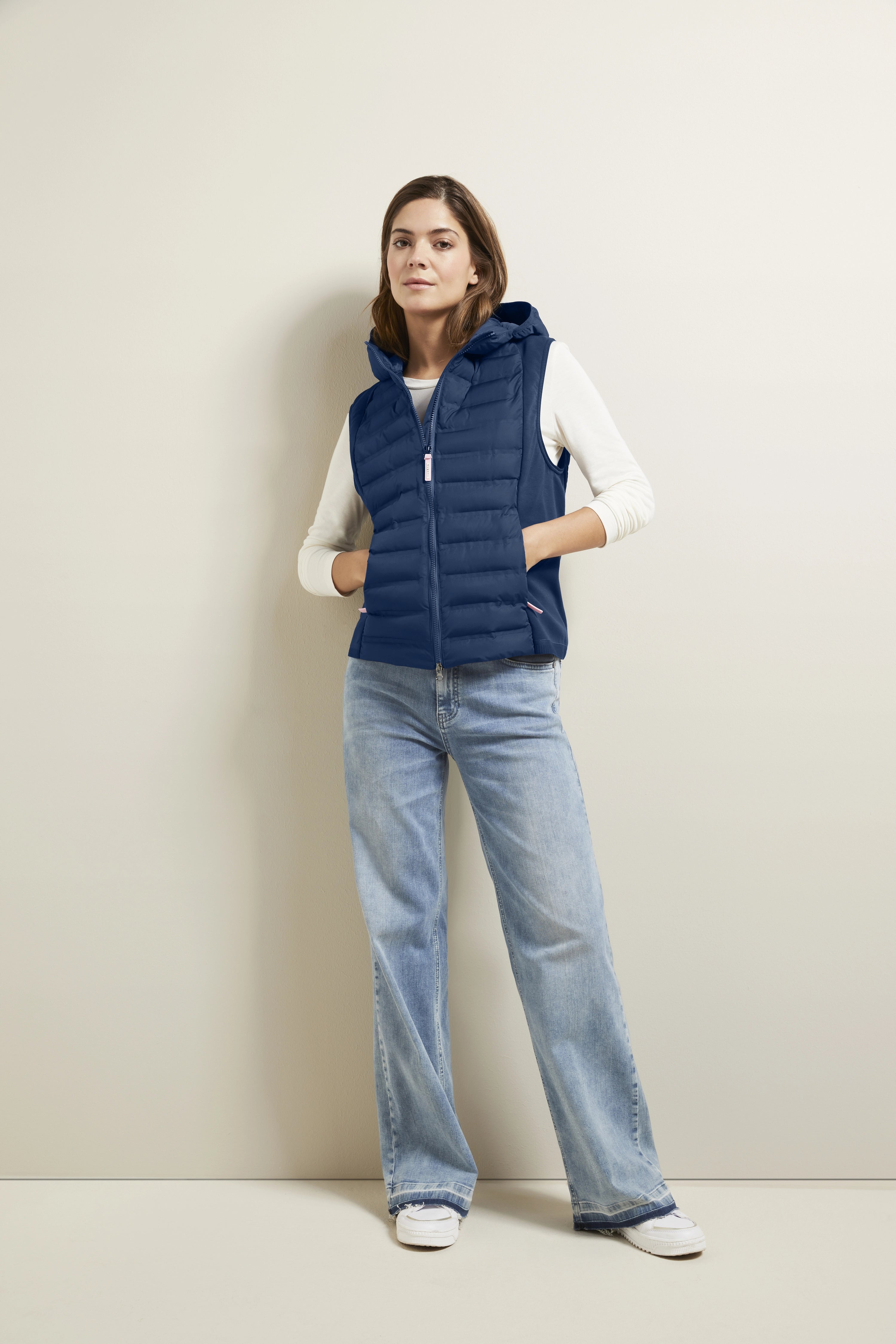 Model draagt blauwe bugatti bodywarmer, wit lang shirt, lichtblauwe jeans. Casual met handen in zakken.