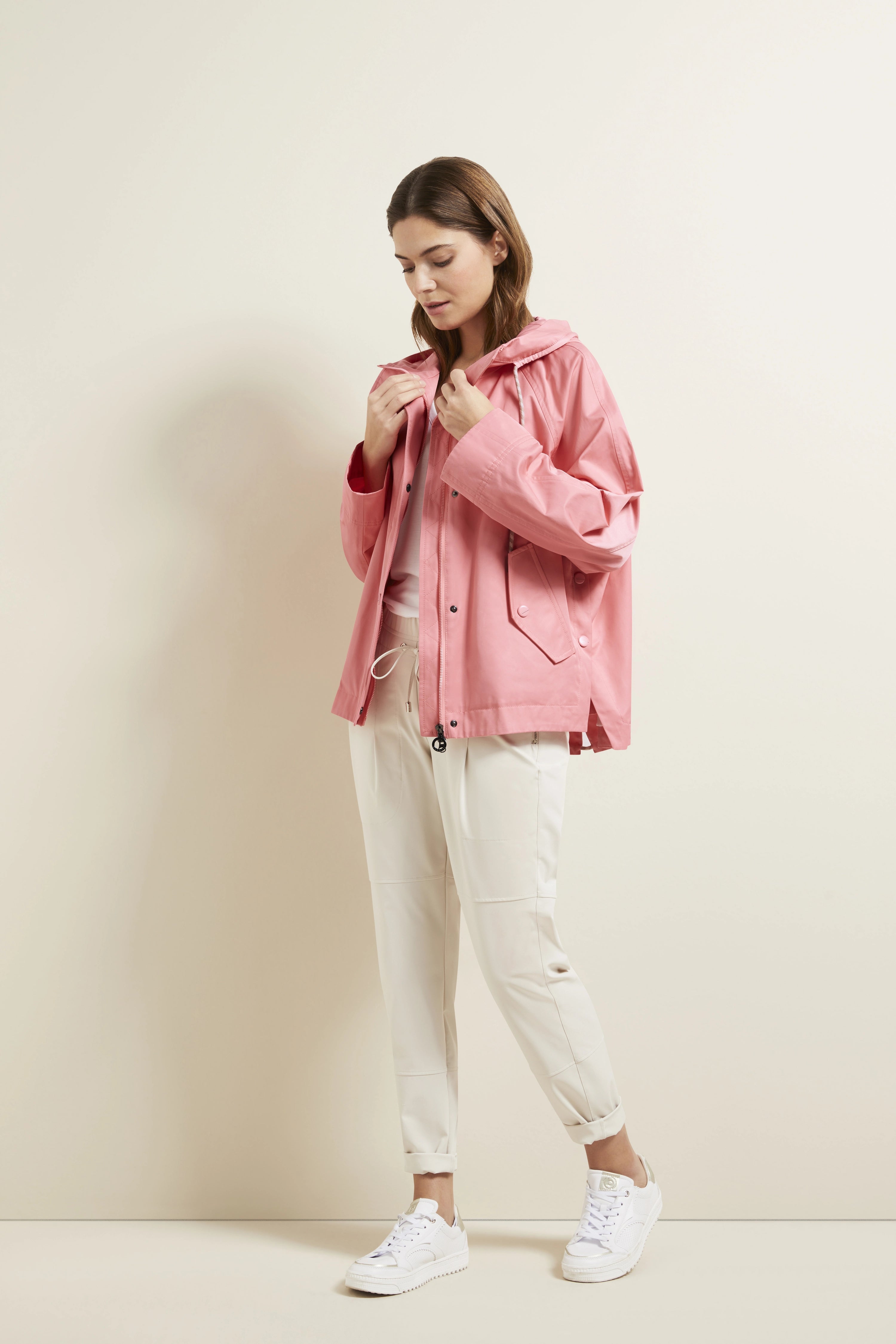 Model in rosa Jacke, weißem Shirt und cremefarbenen Hosen. Lässige Pose, entspanntes Styling.