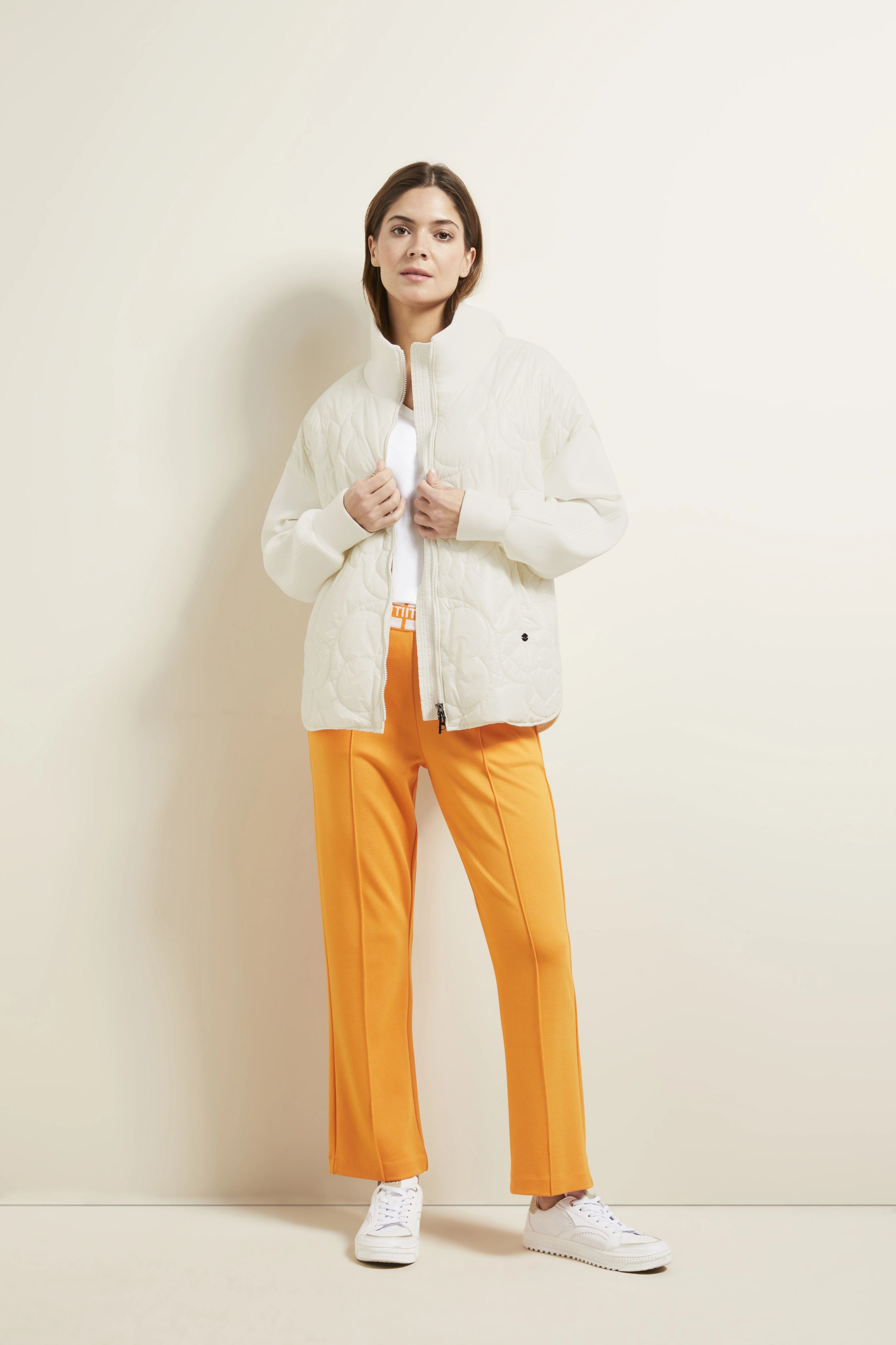 Model trägt cremefarbene Steppjacke, orange Hose, weißes Shirt, weiße Sneaker. Lockeres Posing.