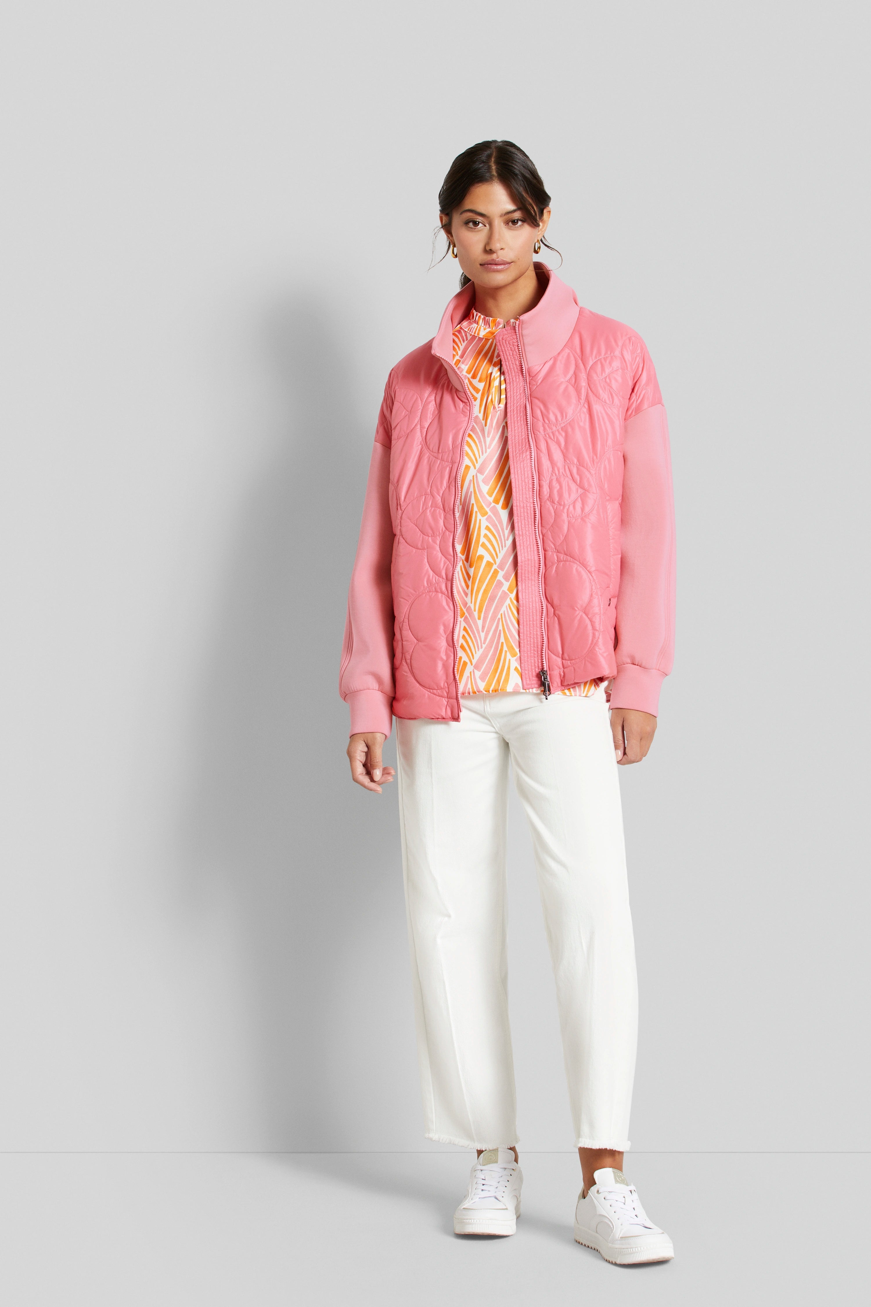 Model in rosa Steppjacke, gemusterter Bluse, weißen Hosen und Sneakers, steht lässig.