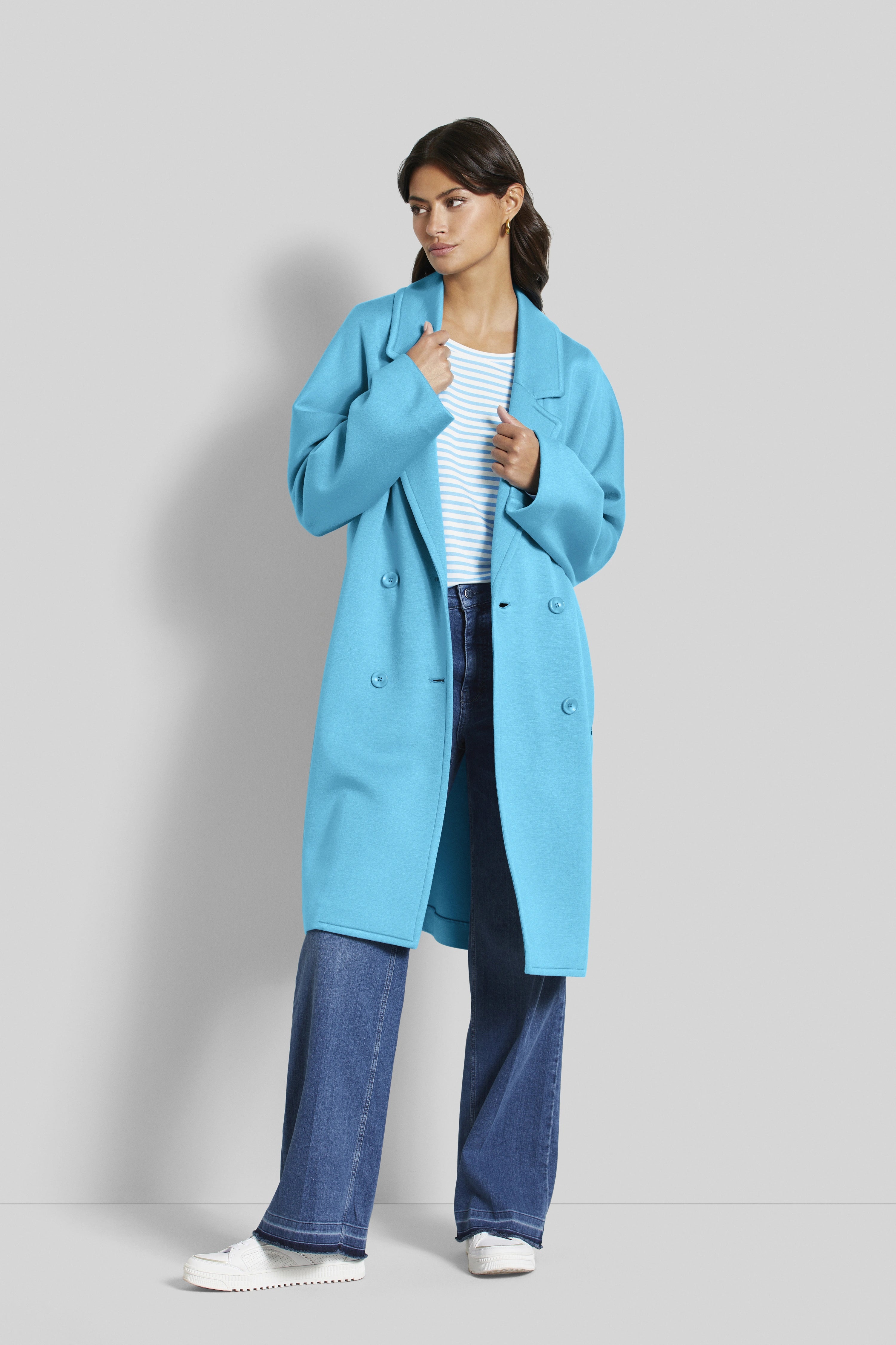 Model in blauem bugatti-Mantel, gestreiftem Shirt, Jeans und weißen Schuhen, lässig stehend.
