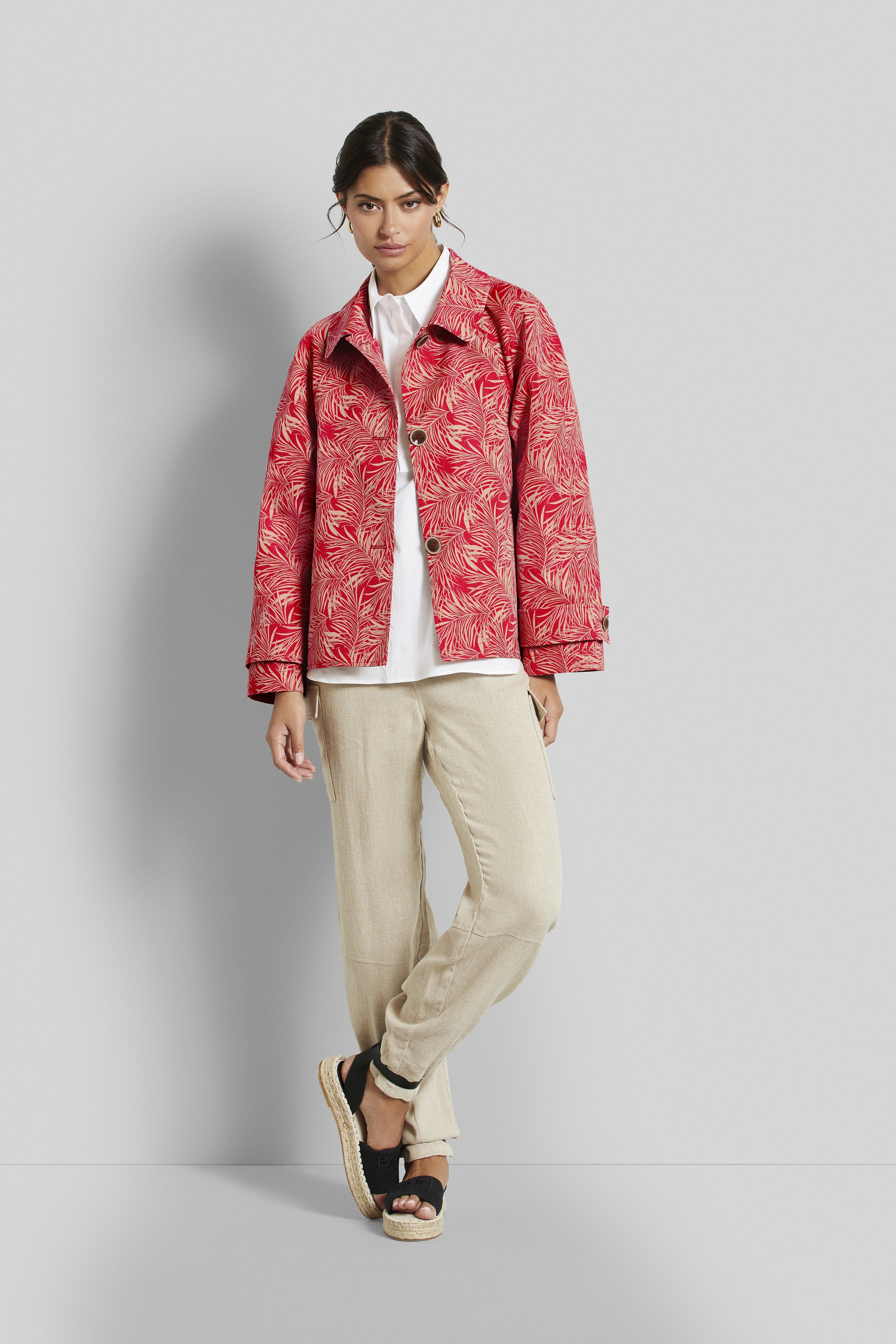 Modelbild mit roter gemusterter Jacke, weißem Hemd, beigefarbener Hose und schwarzen Sandalen. Lässig posierend.