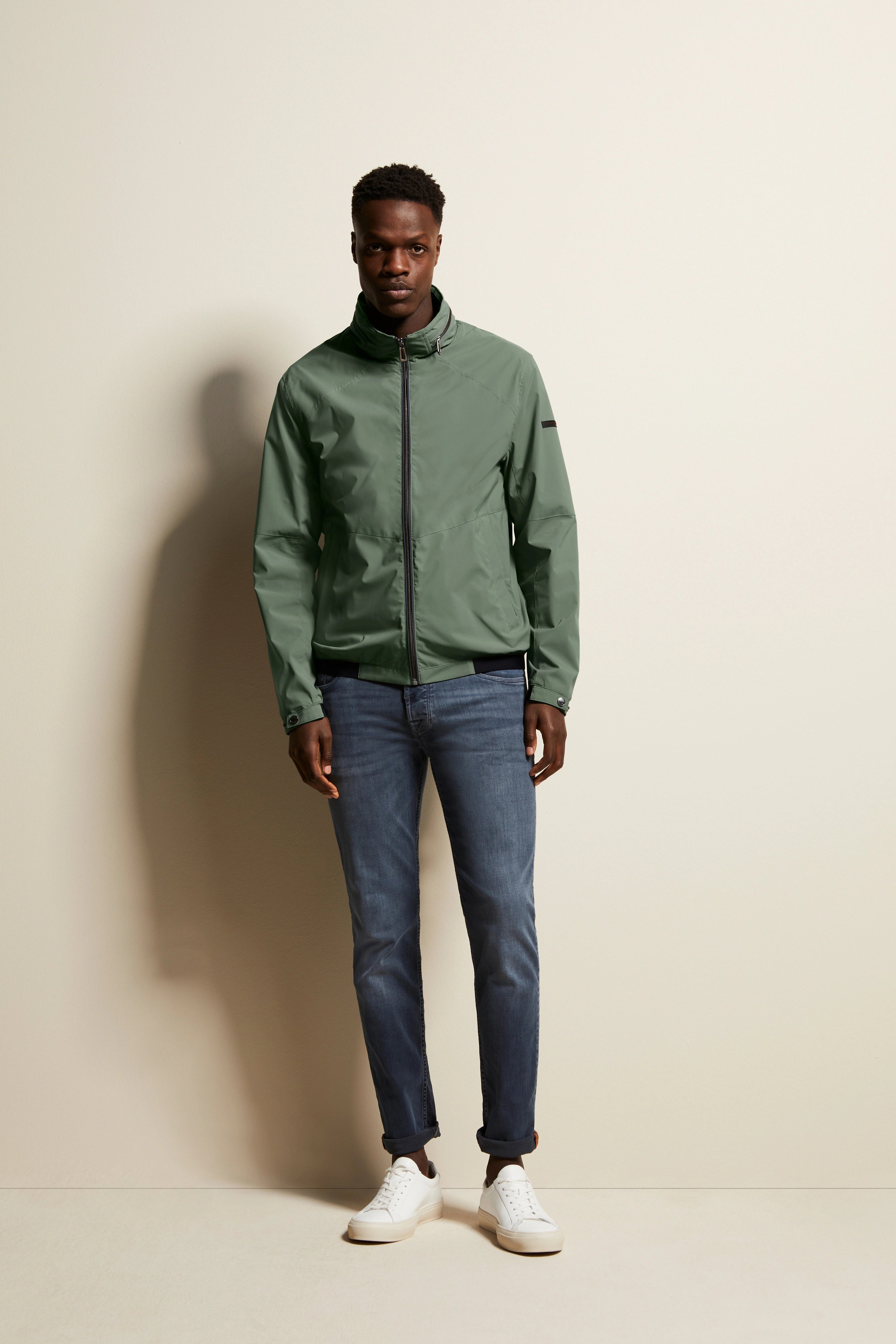 Modelbild mit olivgrüner bugatti Jacke, blauer Jeans, weißen Sneakers, entspannte Pose.