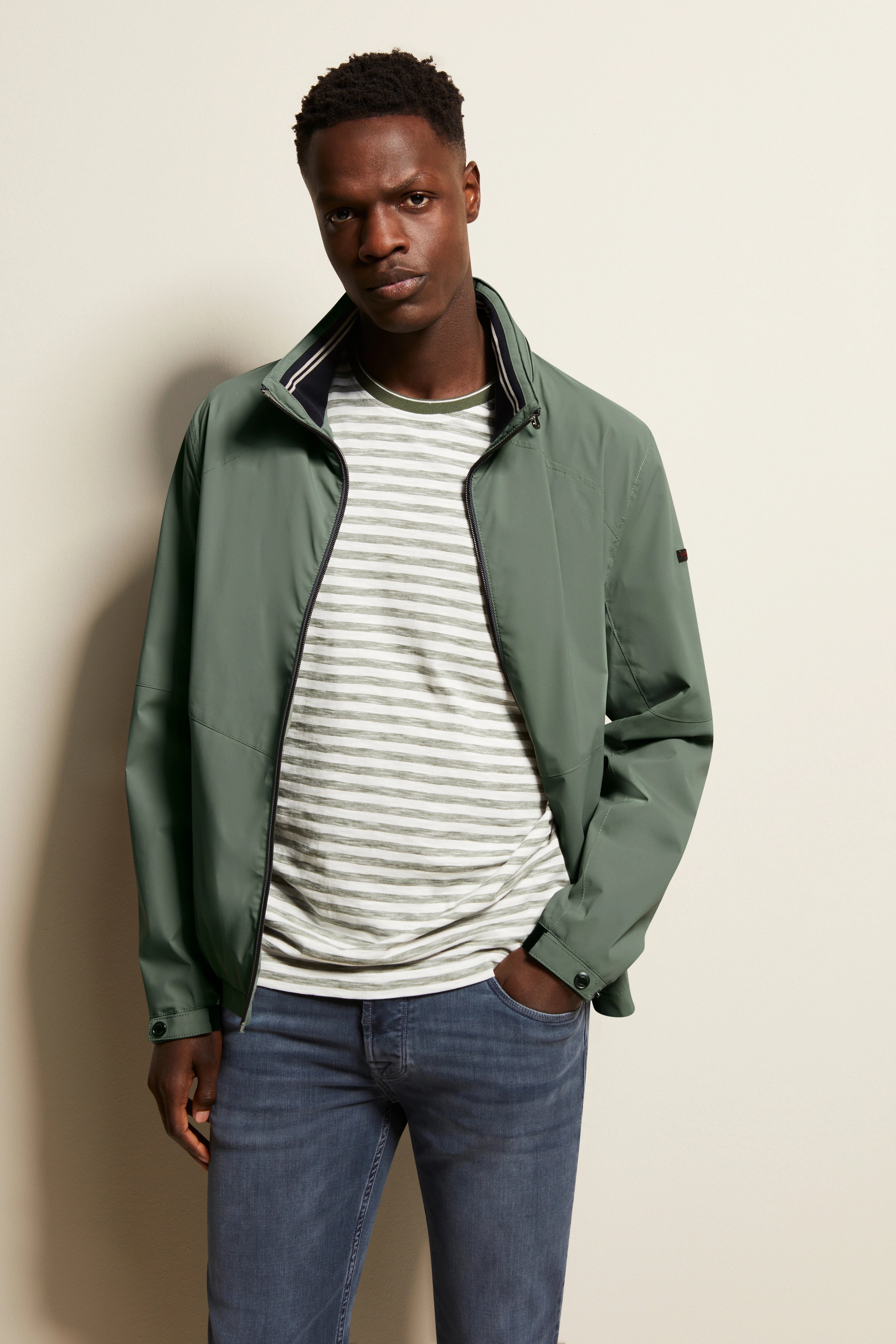 Model in olivgrüner bugatti-Jacke, gestreiftem Shirt und blauer Jeans, lässige Pose an der Wand.