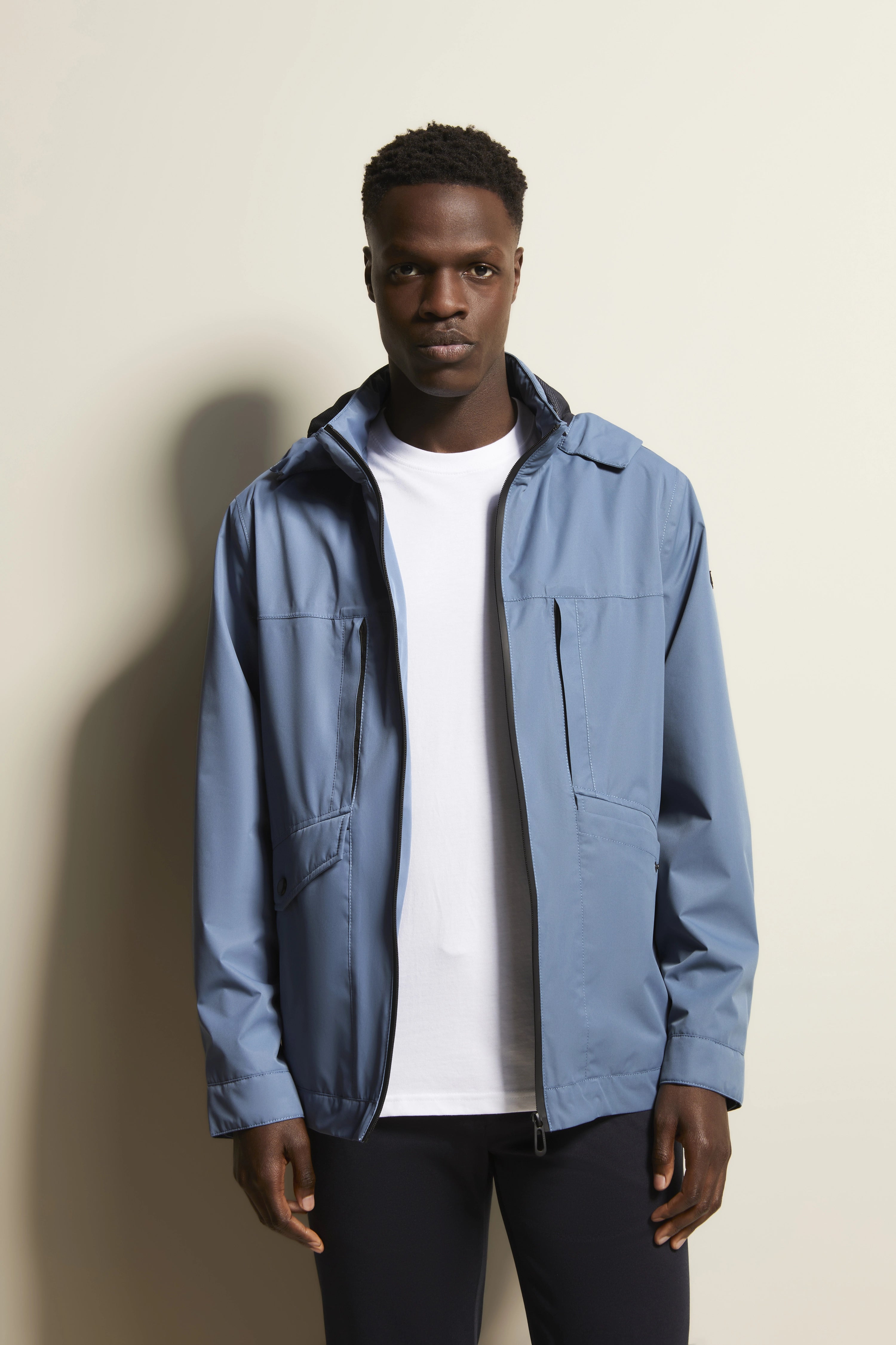 Model in blauwe bugatti-jas, wit T-shirt en donkere broek, staat ontspannen.