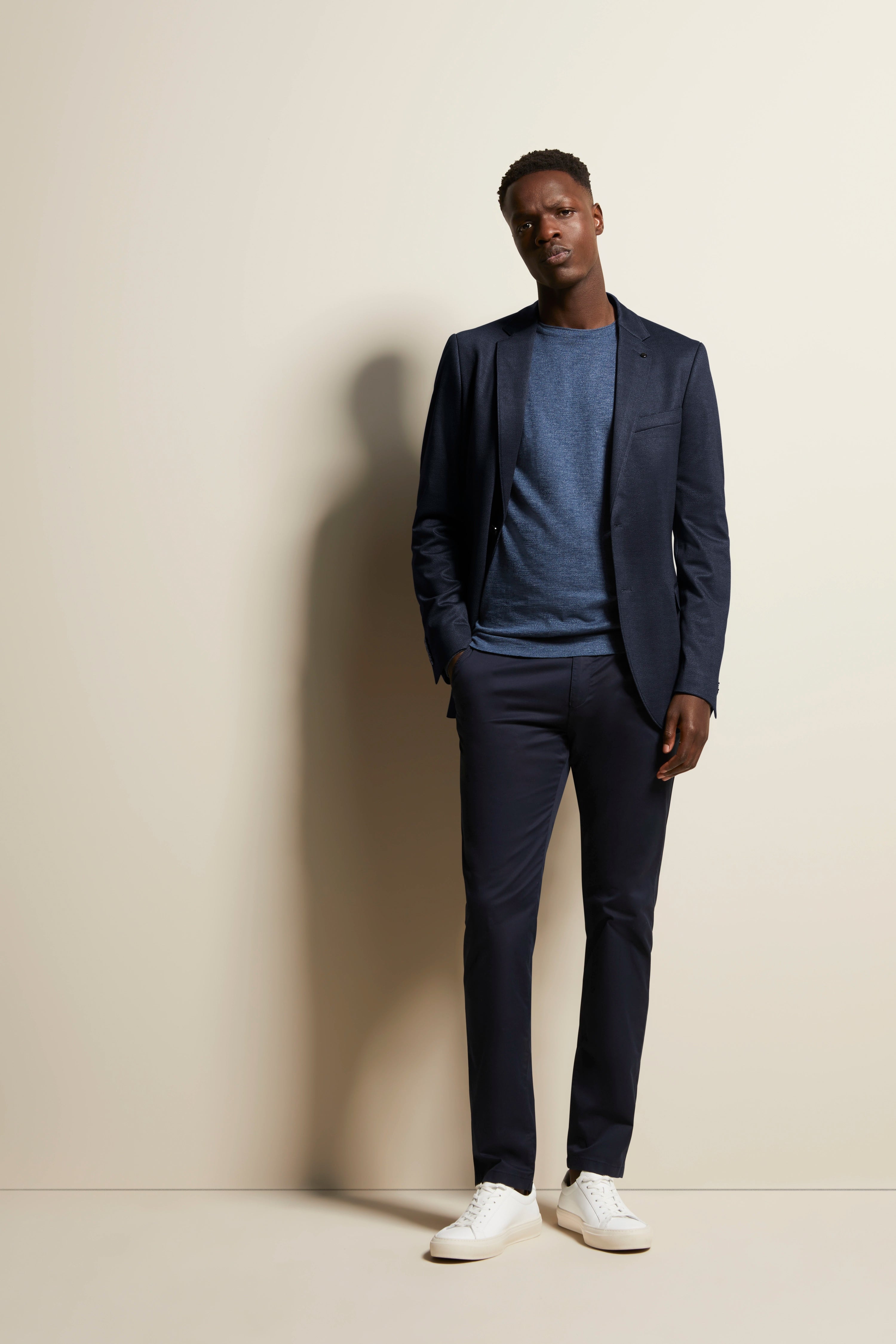 bugatti modelbeeld, man in donkerblauw pak, blauwe trui, witte sneakers, ontspannen pose.