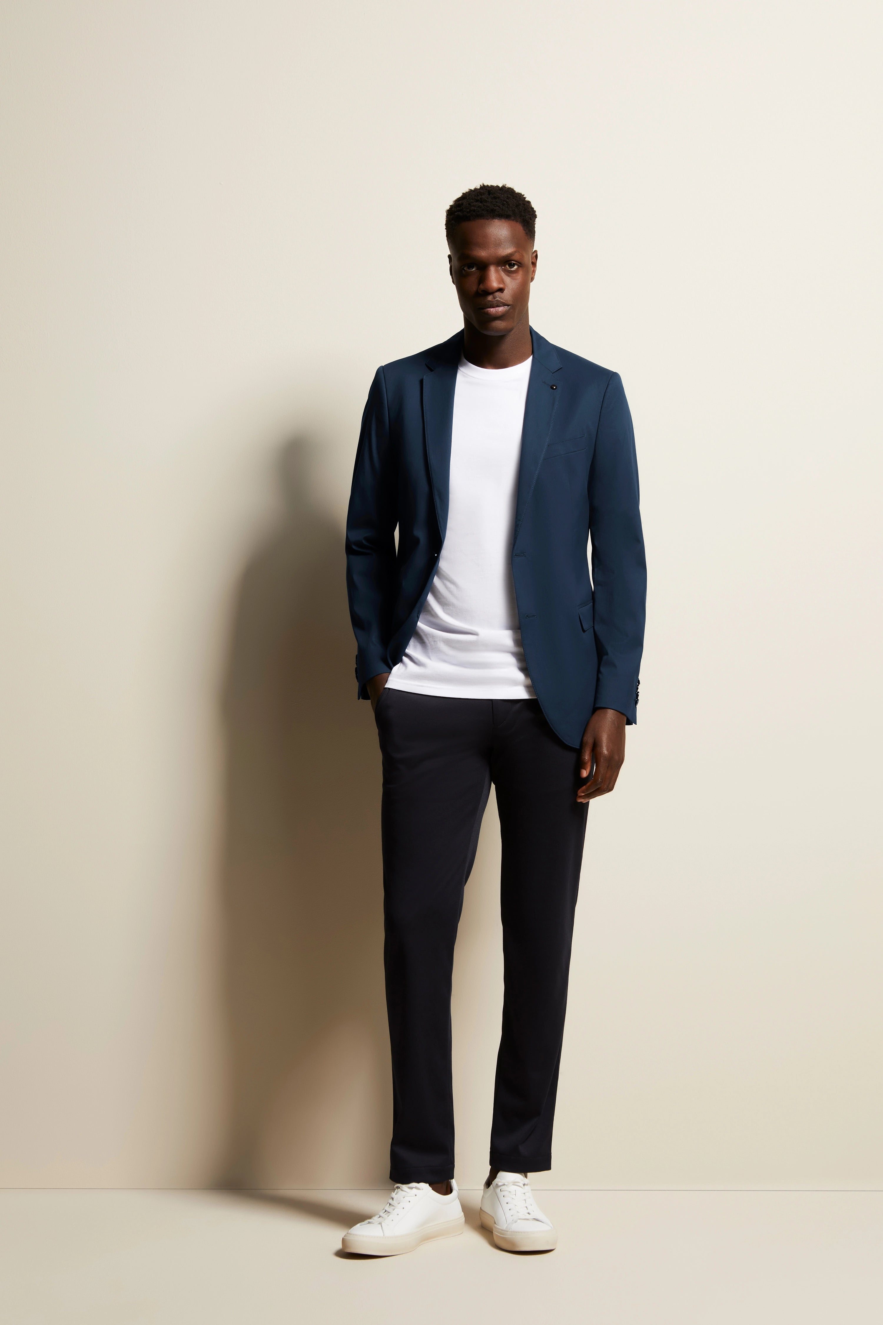 Model in blauwe bugatti blazer, wit T-shirt, donkere broek en witte sneakers, ontspannen pose.