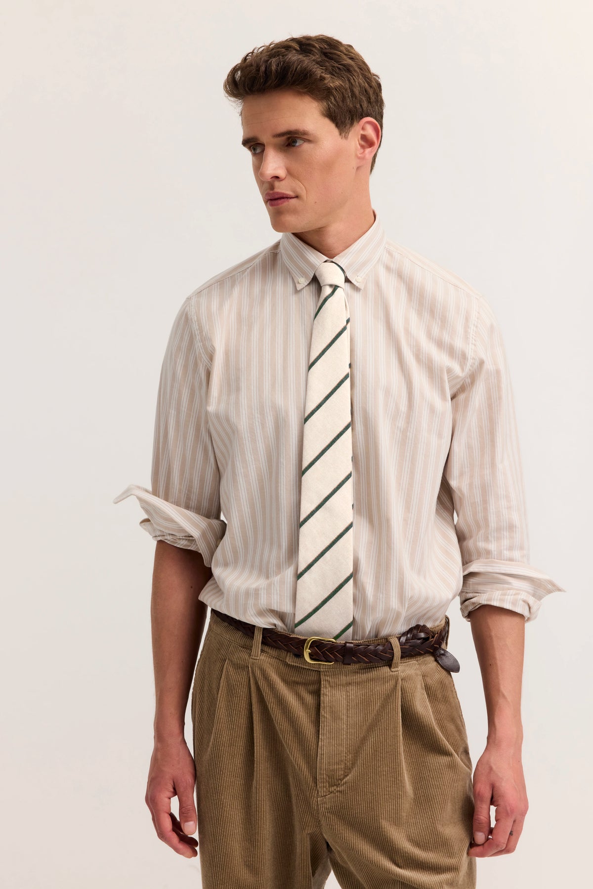 Model in gestreiftem Hemd mit Krawatte, brauner Hose, lässiger Look, seitlicher Blick.