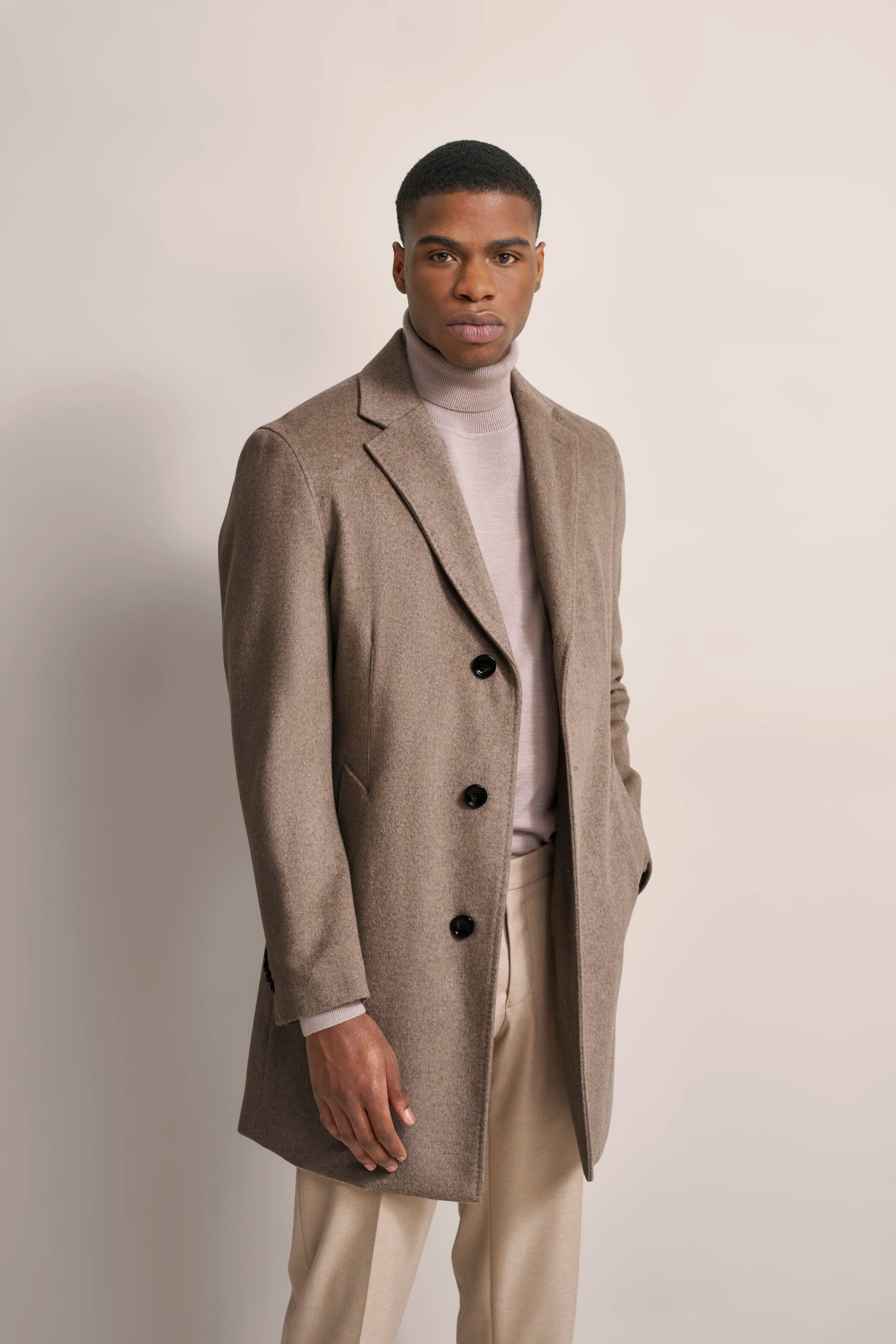 Model in beigem Mantel, Rollkragenpullover und Hose in neutralen Tönen, elegantes, entspanntes Posing.