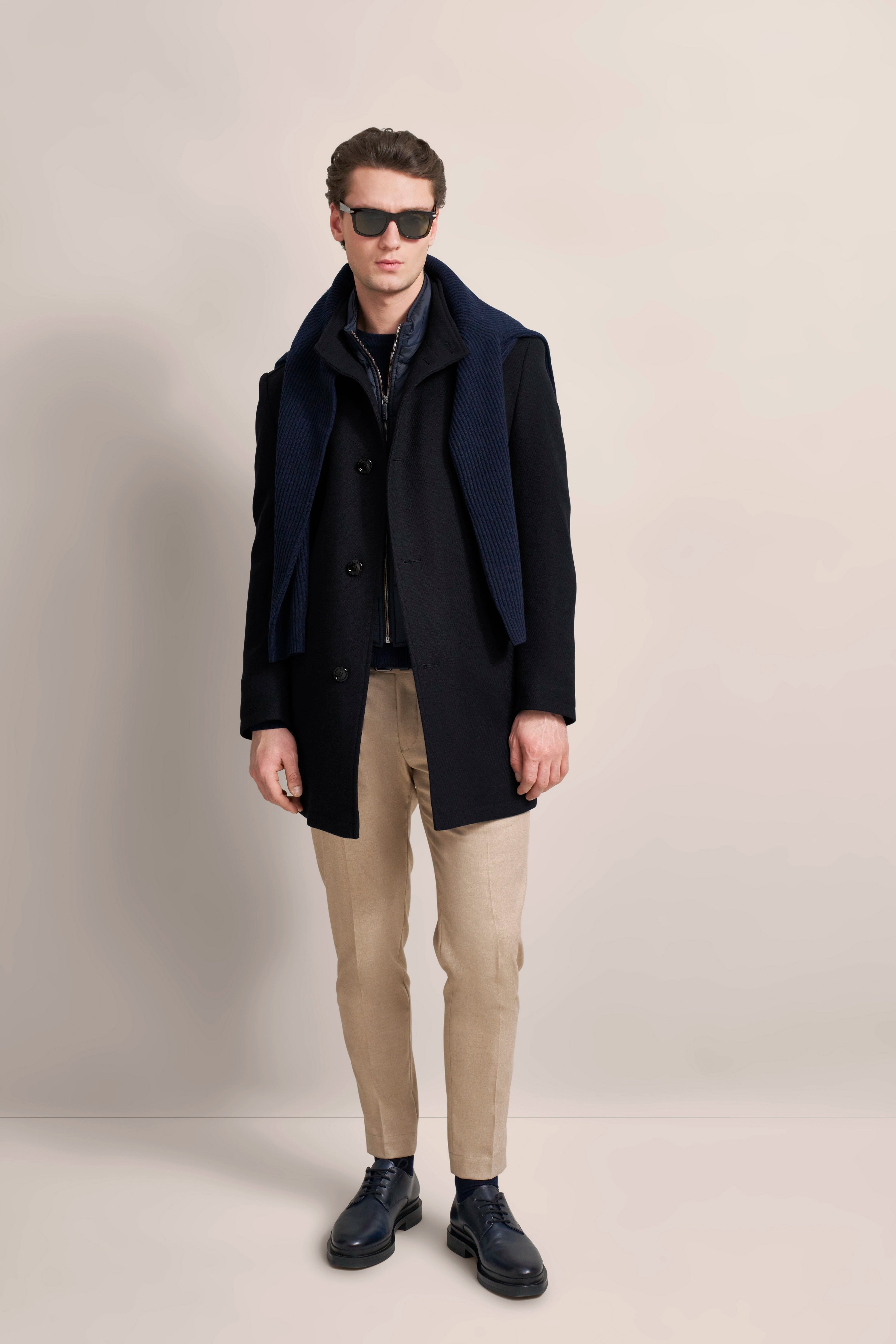 Model in zwarte bugatti-jas met klassieke snit, zijwaartse pose, beige broek.