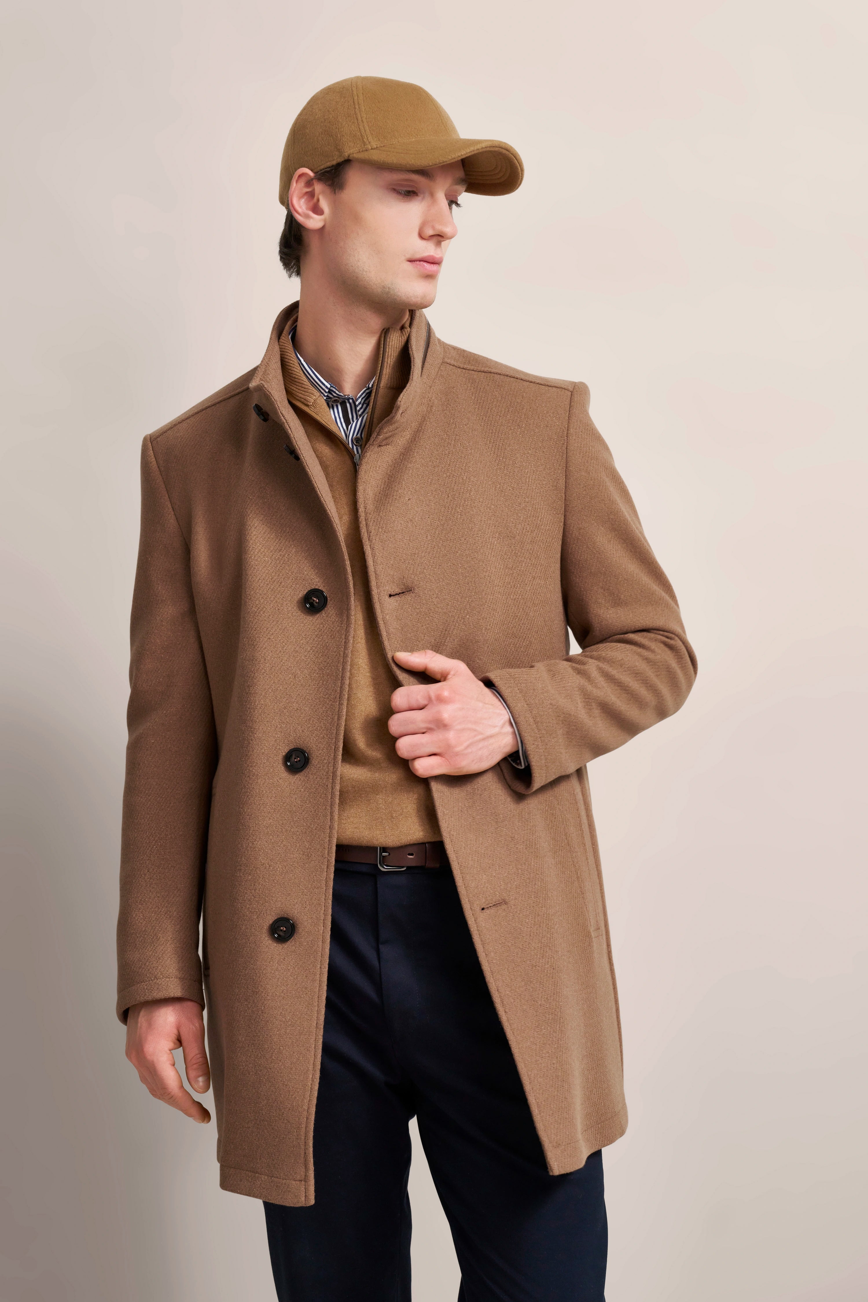 Brauner bugatti-Mantel und Mütze, beigefarbener Pullover, gestreiftes Hemd, Modell in lässiger Pose.