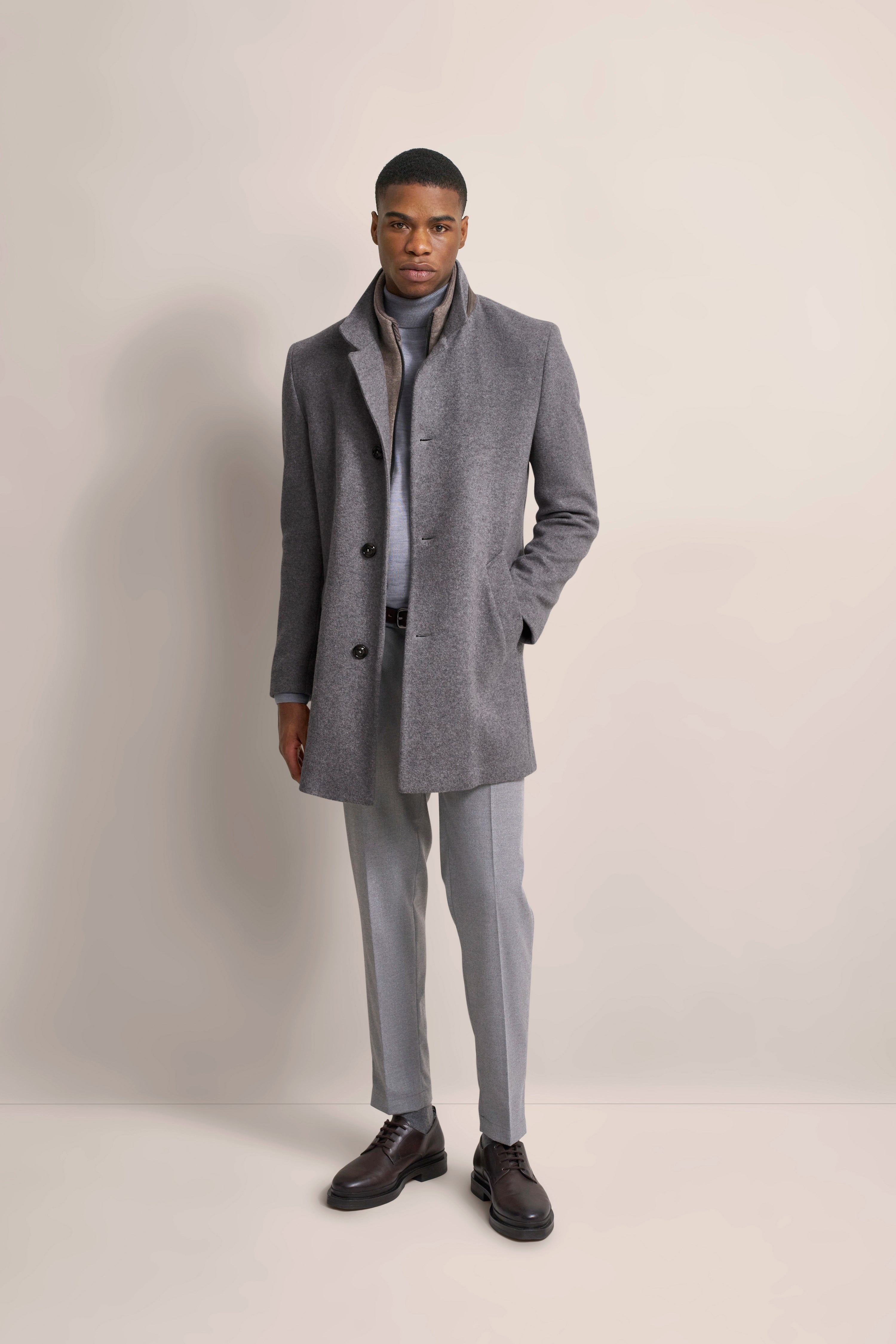 Model in grauem bugatti Mantel, grauem Rollkragen, grauer Hose, schwarze Schuhe, legeres Steh-Posing.