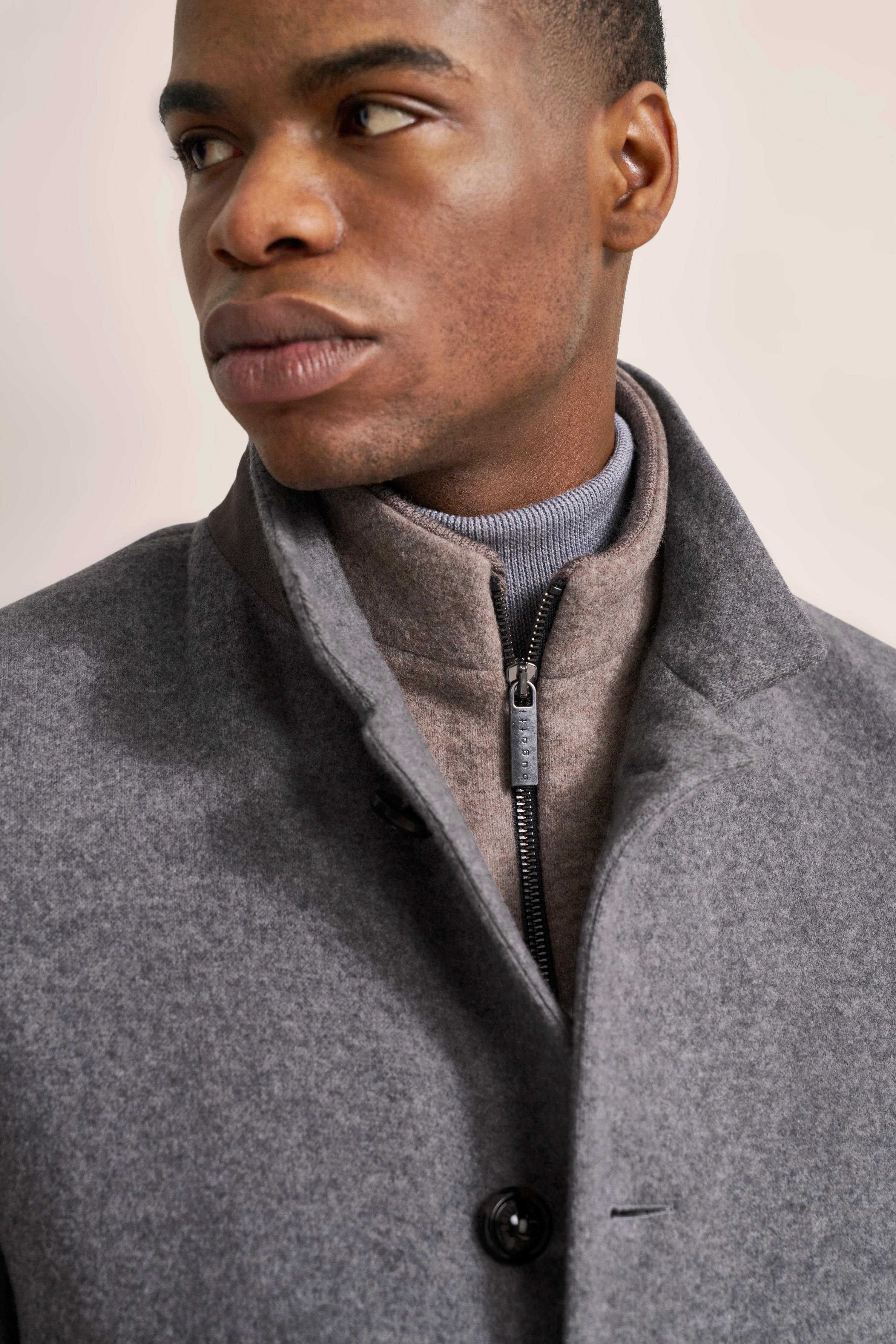 Grauer bugatti Mantel und beiger Rollkragenpullover, Modelansicht mit ruhigem, nachdenklichem Ausdruck.