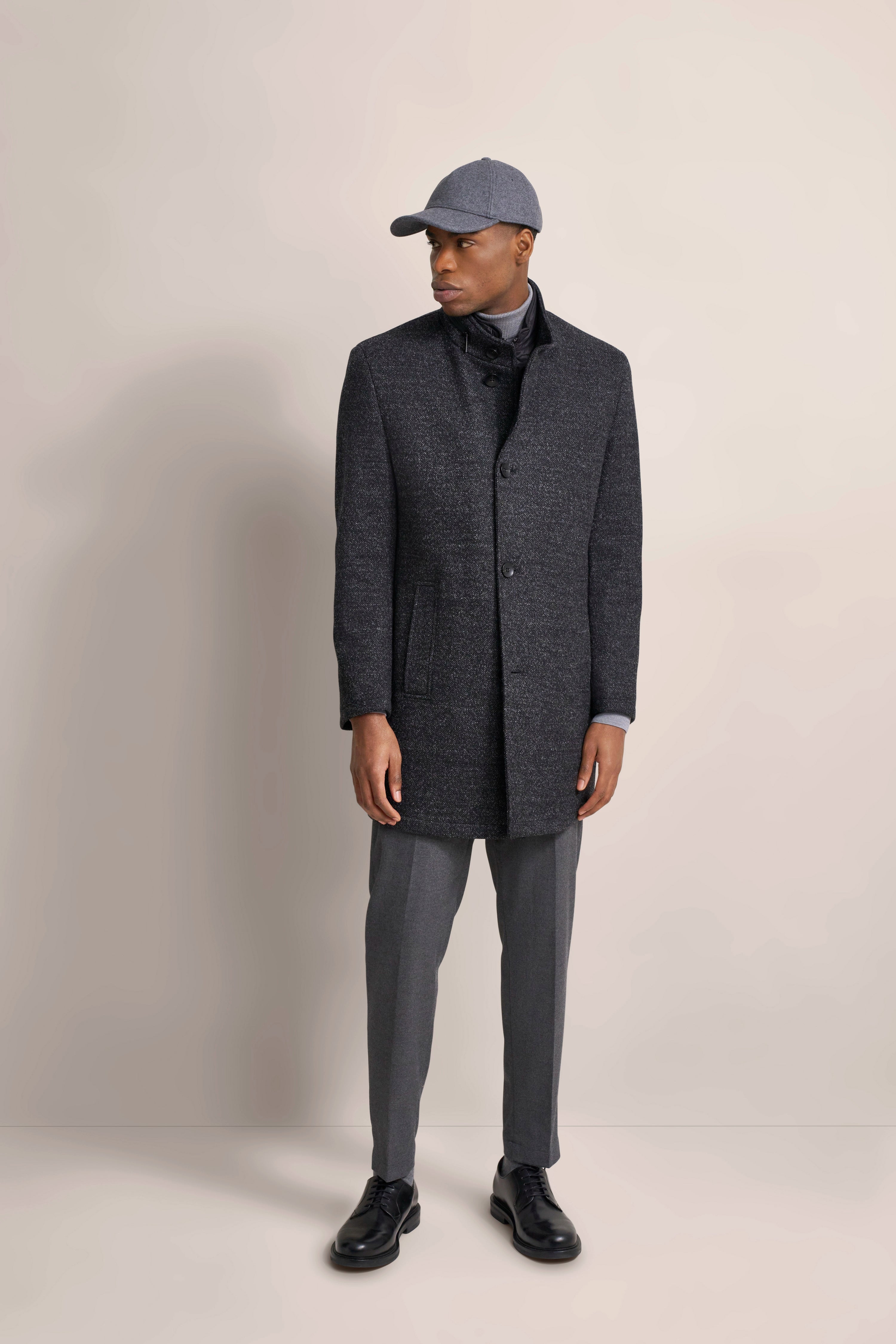 Männermodell in grauem bugatti Mantel, grauer Hose und Basecap. Elegante Pose, seitlich blickend.