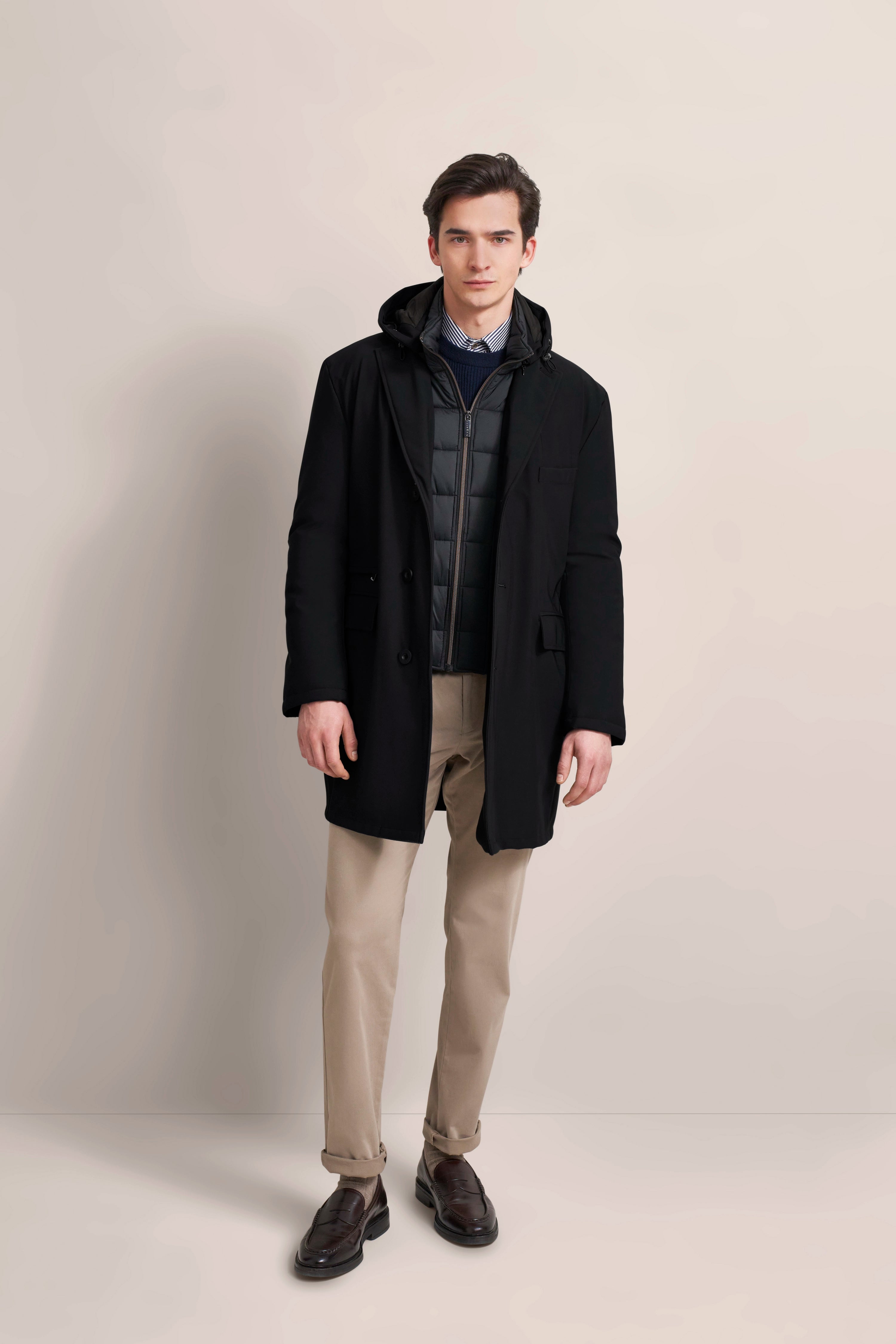 bugatti Model in schwarzem Mantel, gestepptem Hoodie, beiger Hose und braunen Loafers, entspanntes Posing.