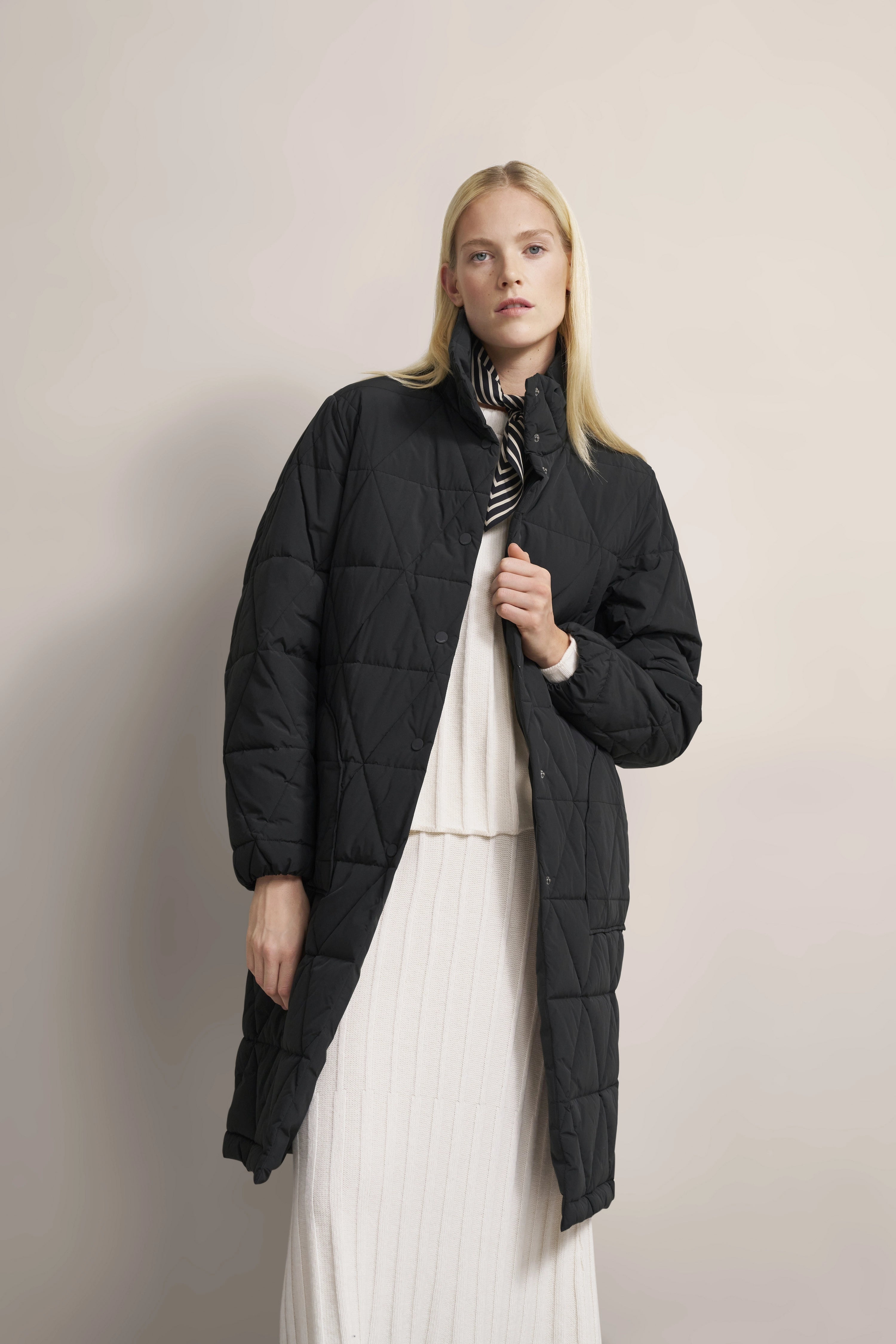 Model trägt schwarzen bugatti Steppmantel, weiße Strickkleid, schwarz-weißes Halstuch. Entspanntes Posing.