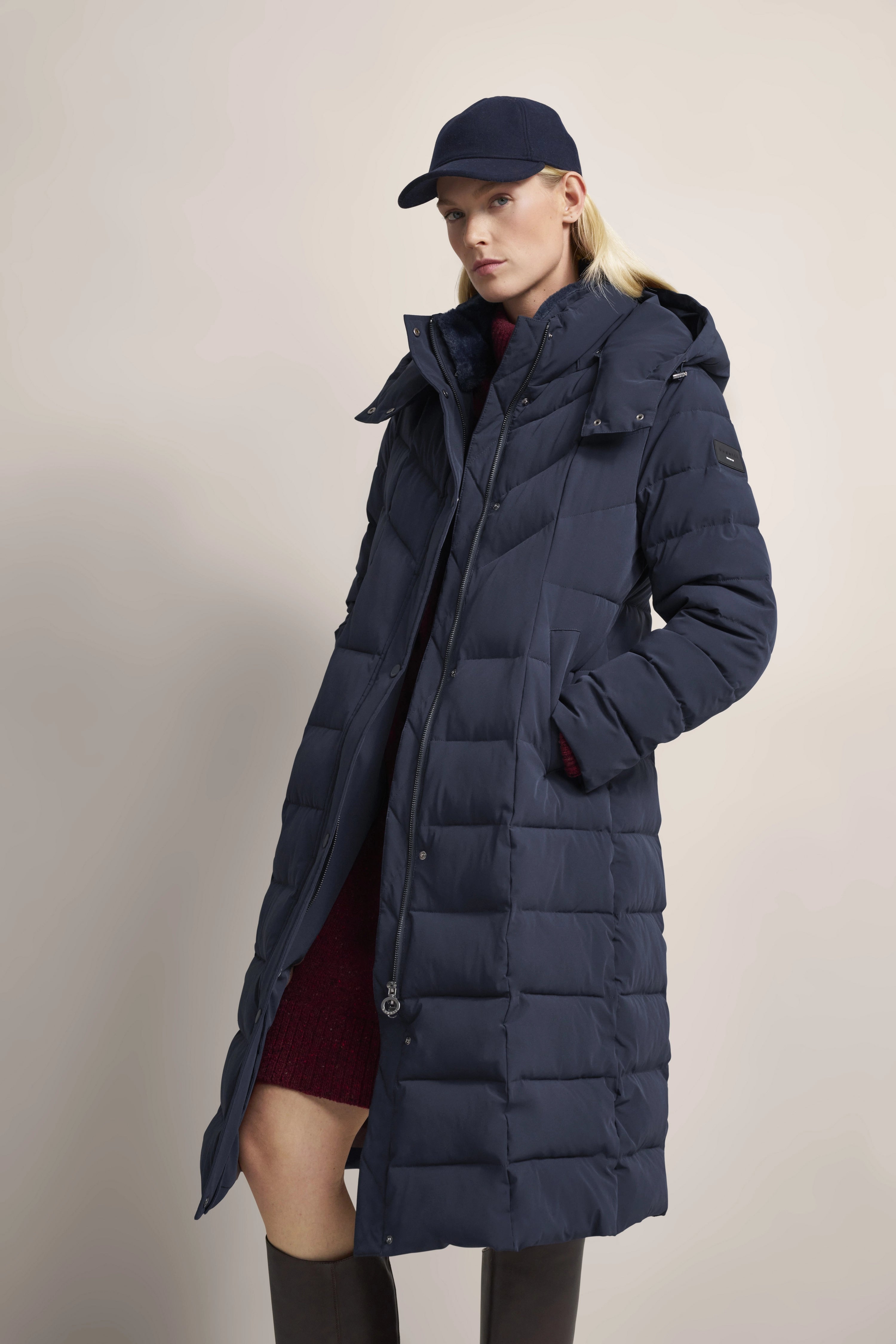 Model in navy Steppmantel, burgunderrotem Kleid, schwarzer Mütze und Stiefeln. Lässige Pose.