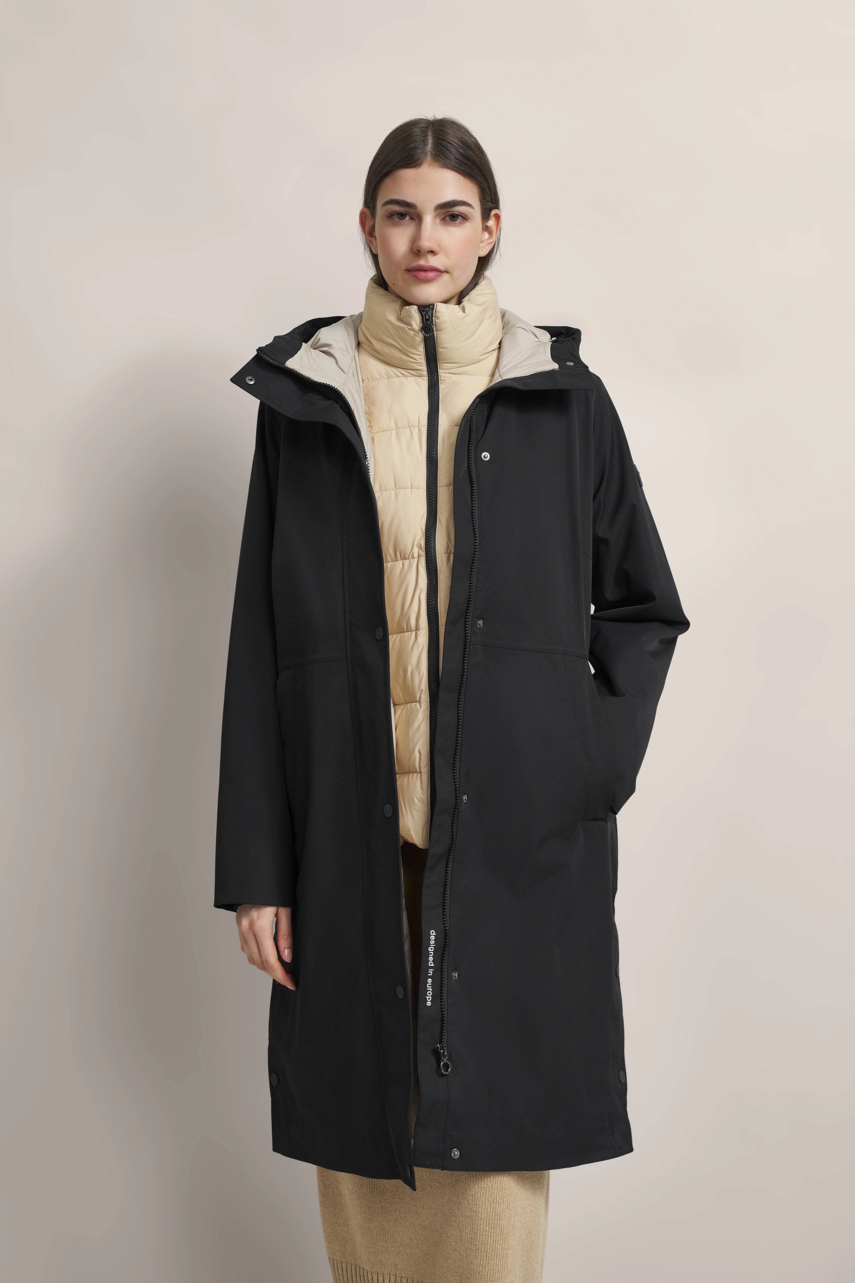 Dame in schwarzem bugatti-Mantel über beigem Steppjacke, entspannte Pose.