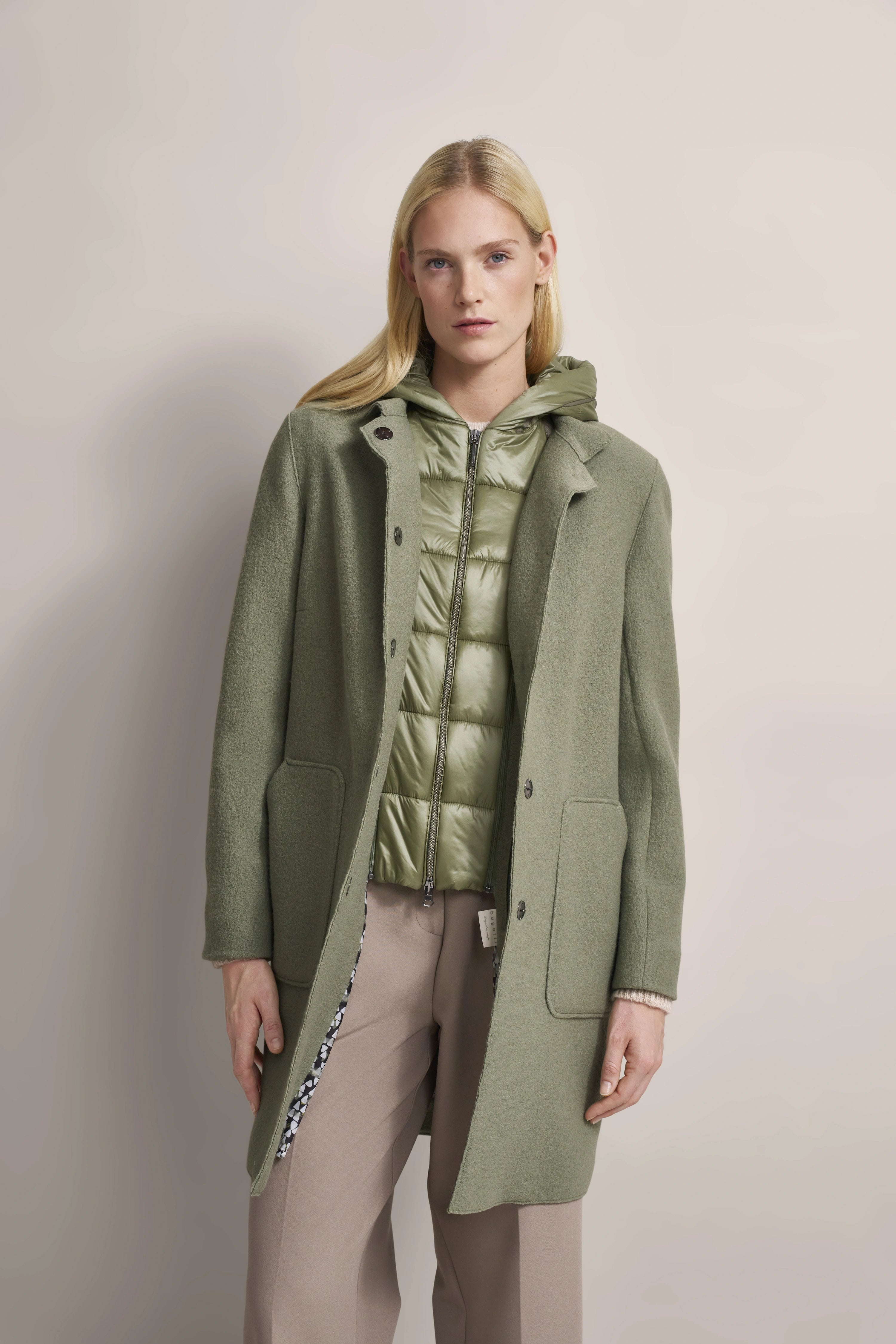 Model in olivgrünem bugatti Mantel und Steppjacke, beige Hose, entspanntes Posing.