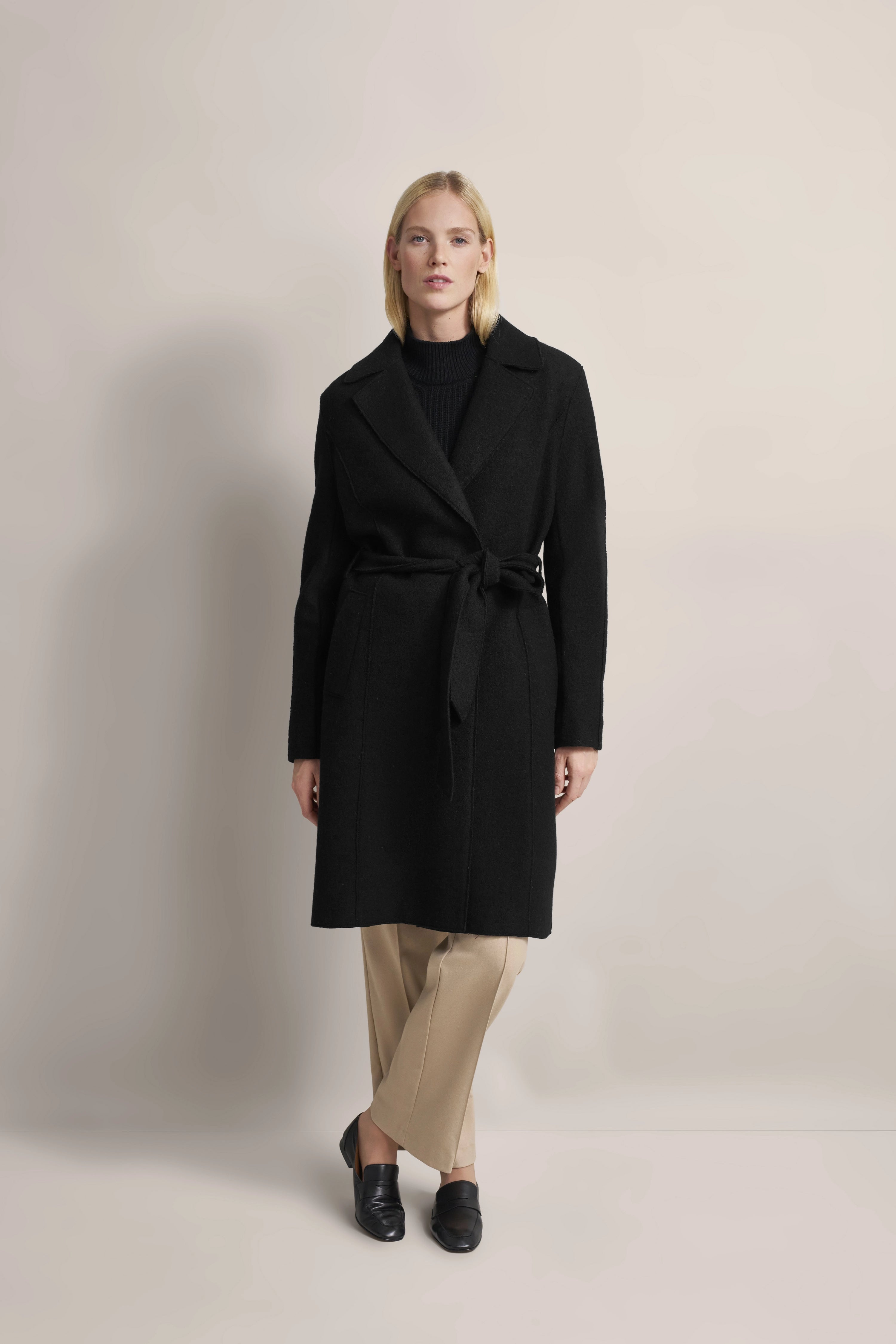 Schwarzer bugatti Mantel, beiger Hose, schwarzem Pullover. Modelbild, entspannt stehend.