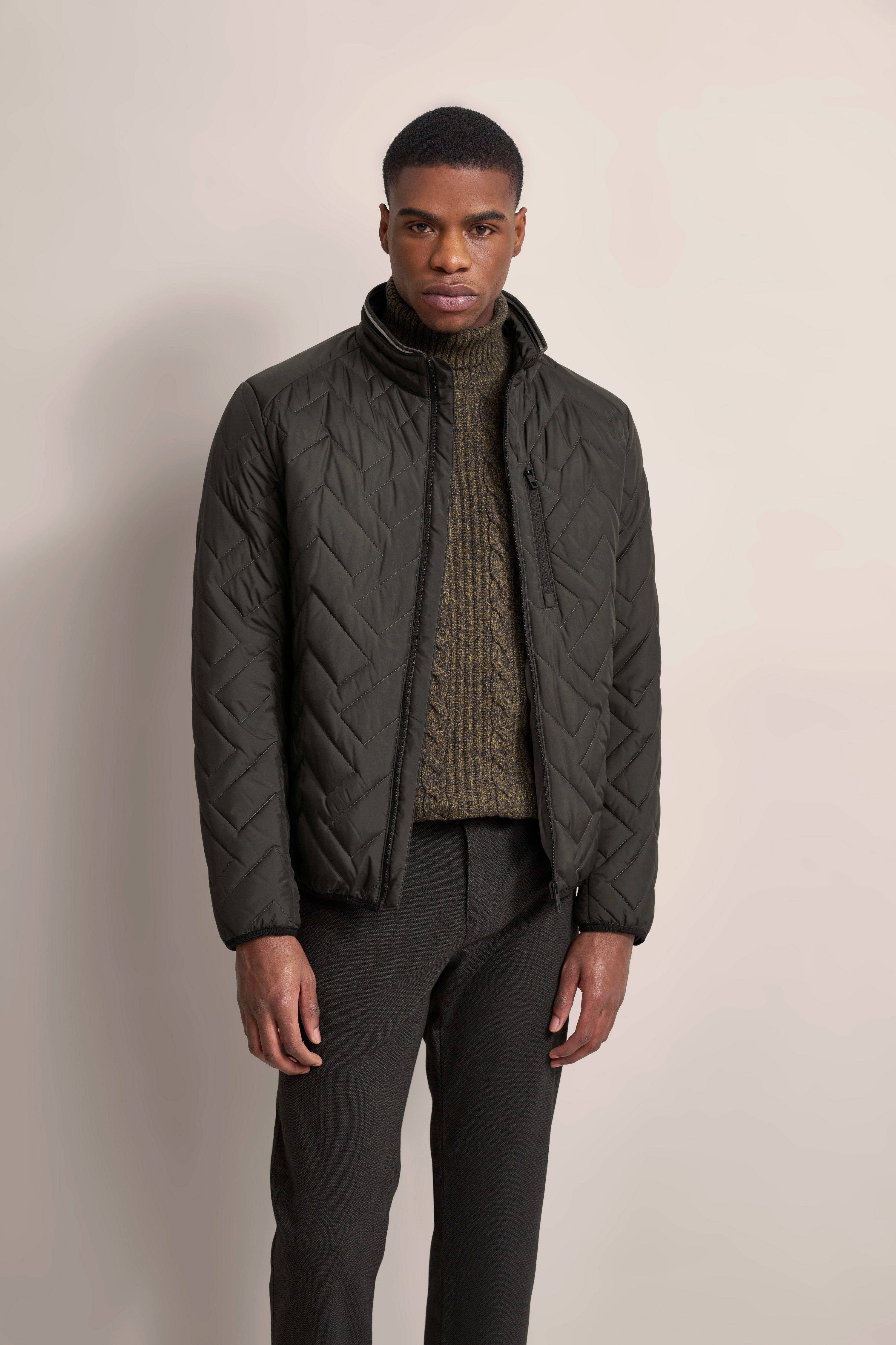 Model mit schwarzer Steppjacke, olivgrünem Strickpullover und schwarzen Hosen. Entspannte Pose.