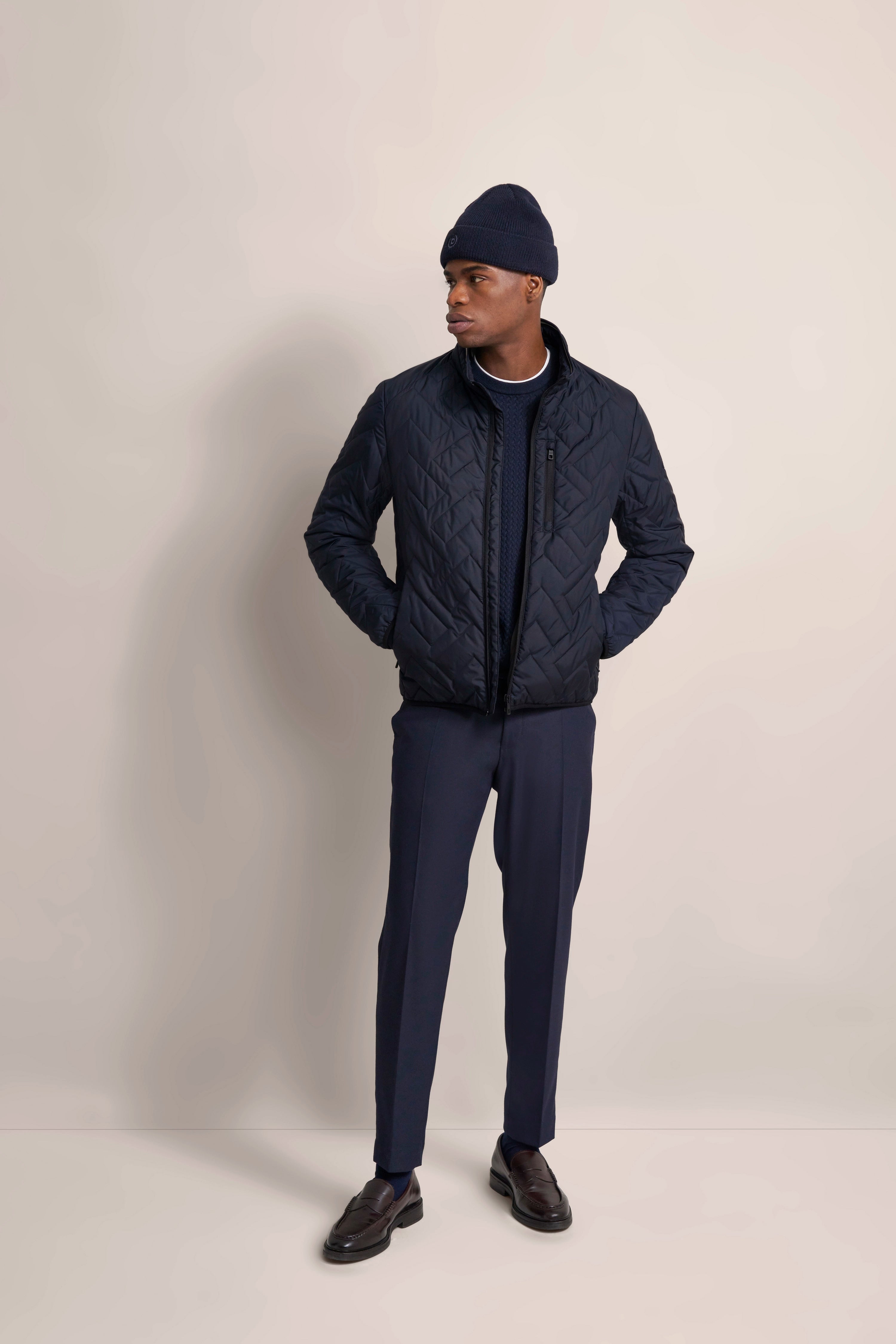 Model in dunkelblauer Steppjacke, Mütze, Hose und Pullover von bugatti, legeres Posing.