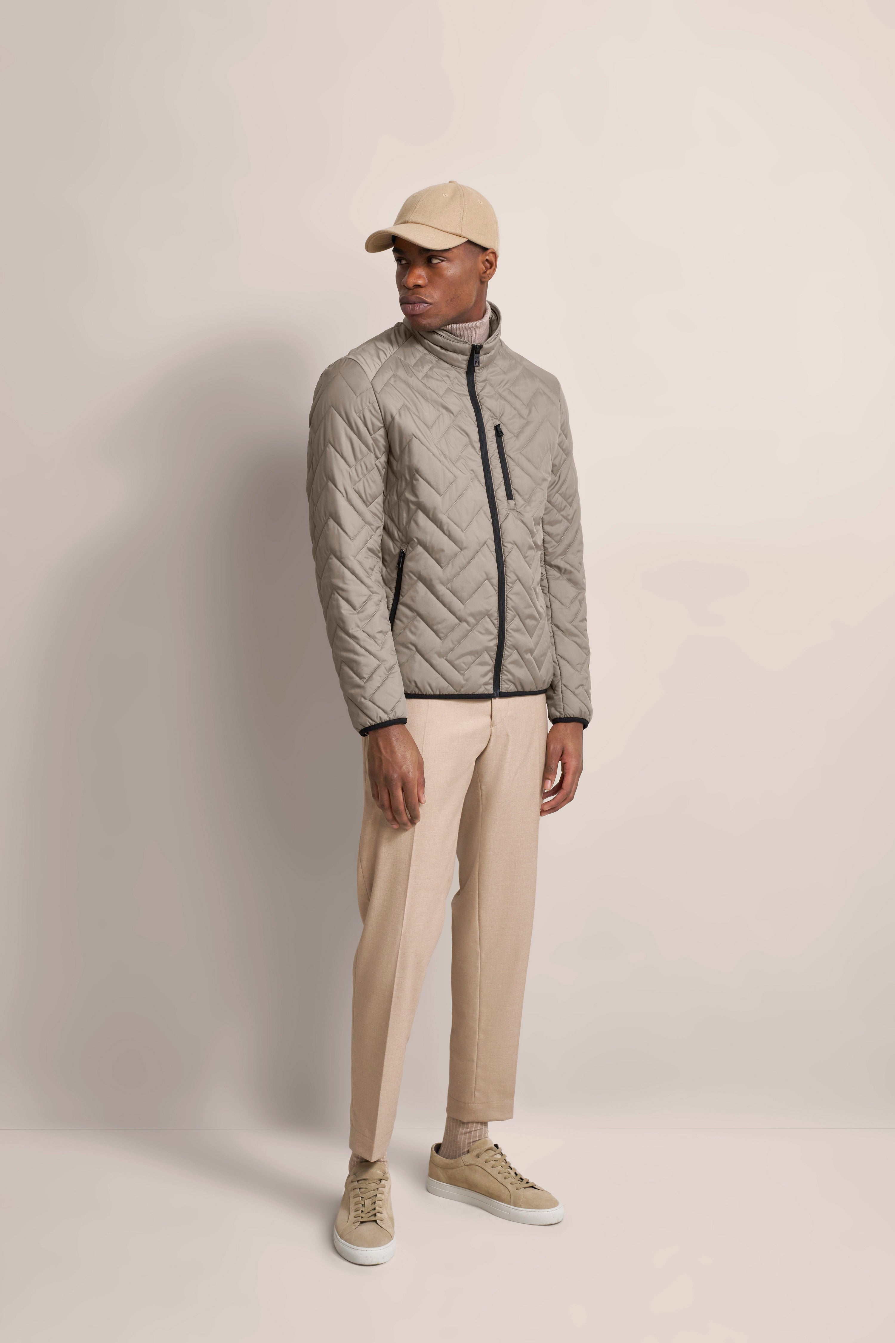 Model in taupe Steppjacke, beige Hose und Cap, beige Schuhe. Lässige Pose seitlich. bugatti Stil.