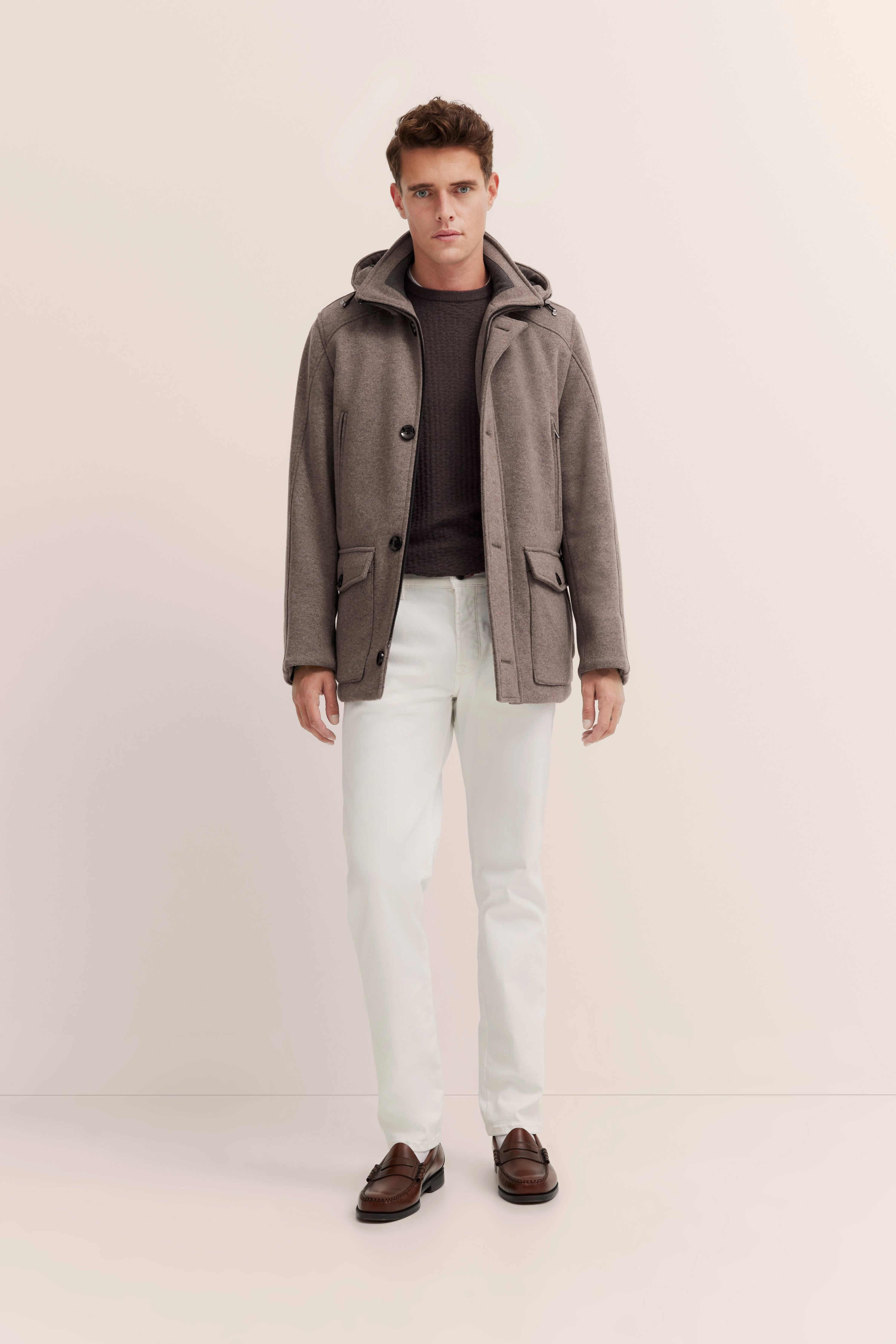Model in brauner bugatti Jacke, dunklem Pullover, weißen Hosen, braunen Loafern, entspannte Haltung.
