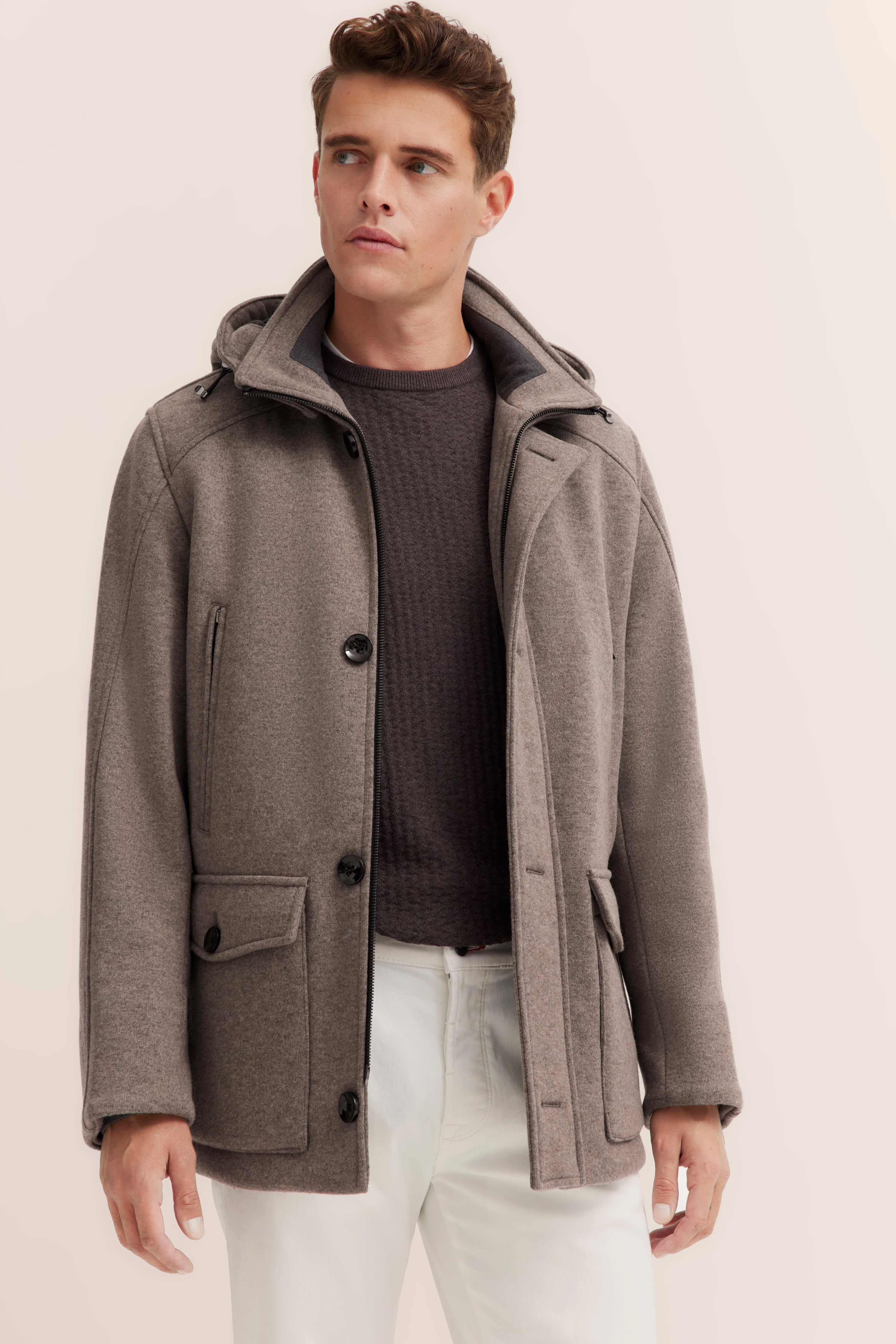 bugatti Mantel in Taupe mit Kapuze, dunkler Pullover, weiße Hose, Modelbild, entspannte Pose.