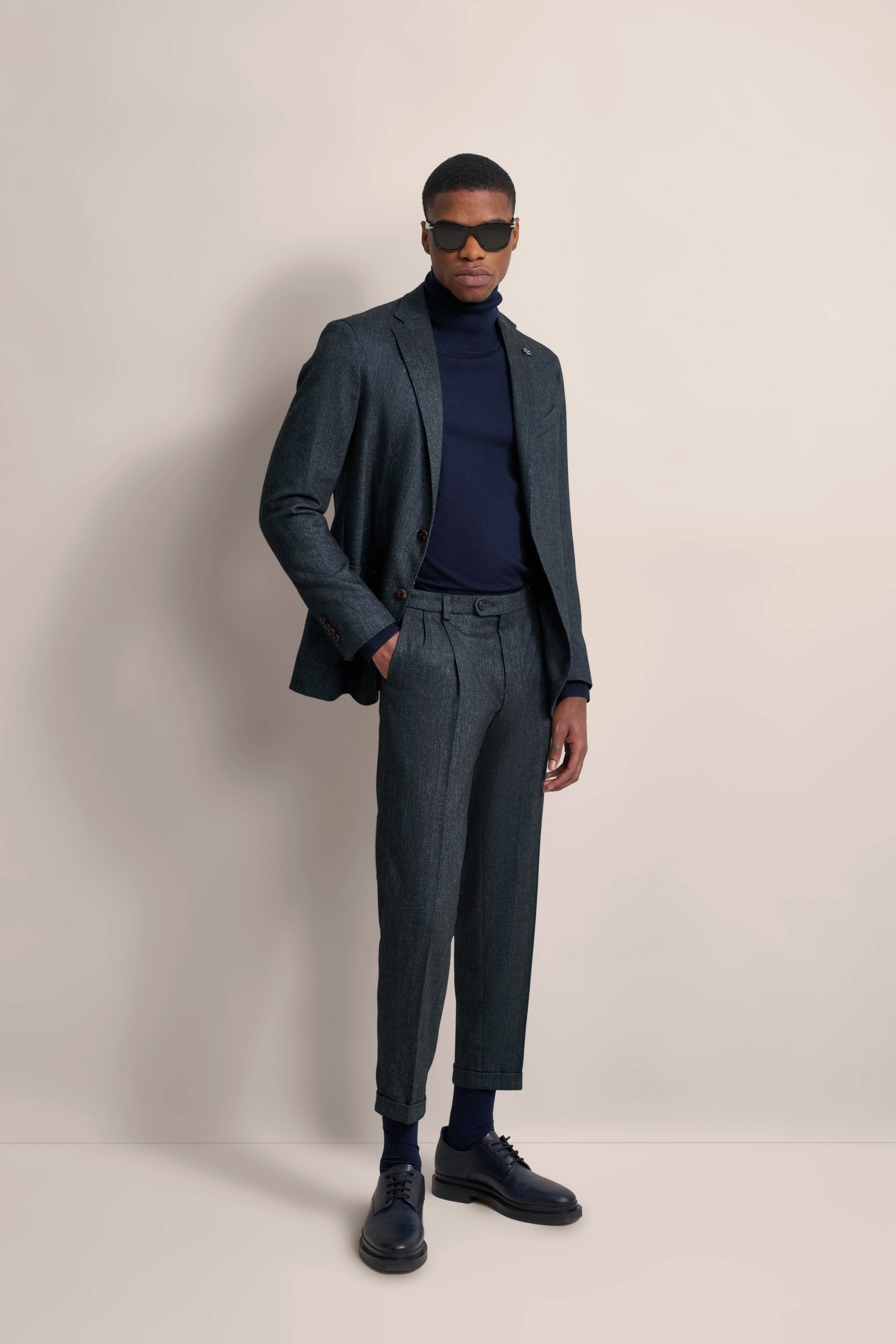 Model in grijs pak met blauwe coltrui, ontspannen pose, bugatti stijl en elegantie.