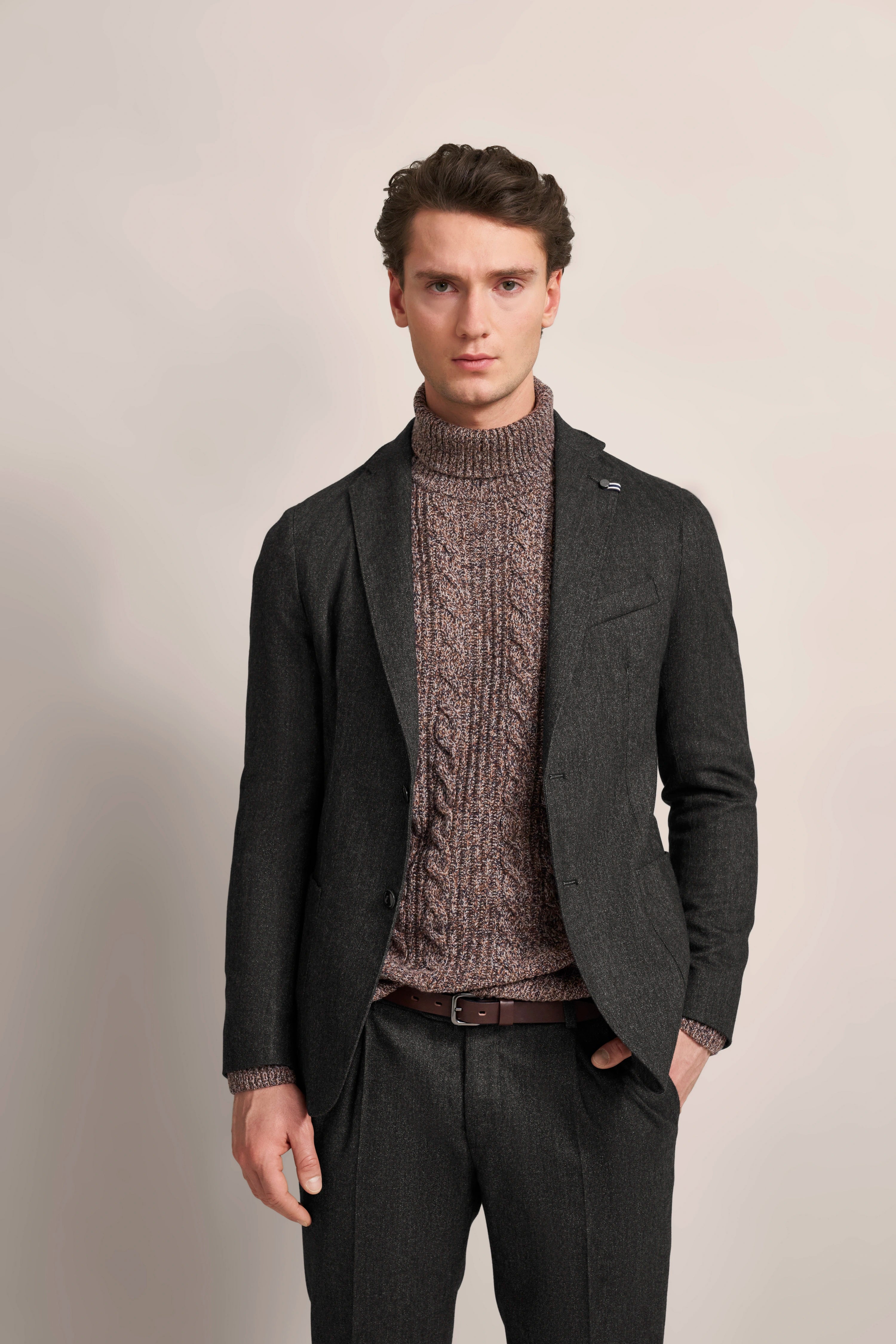 Elegant model shot: Gray bugatti suit, brown sweater, relaxed pose, hands in pockets.