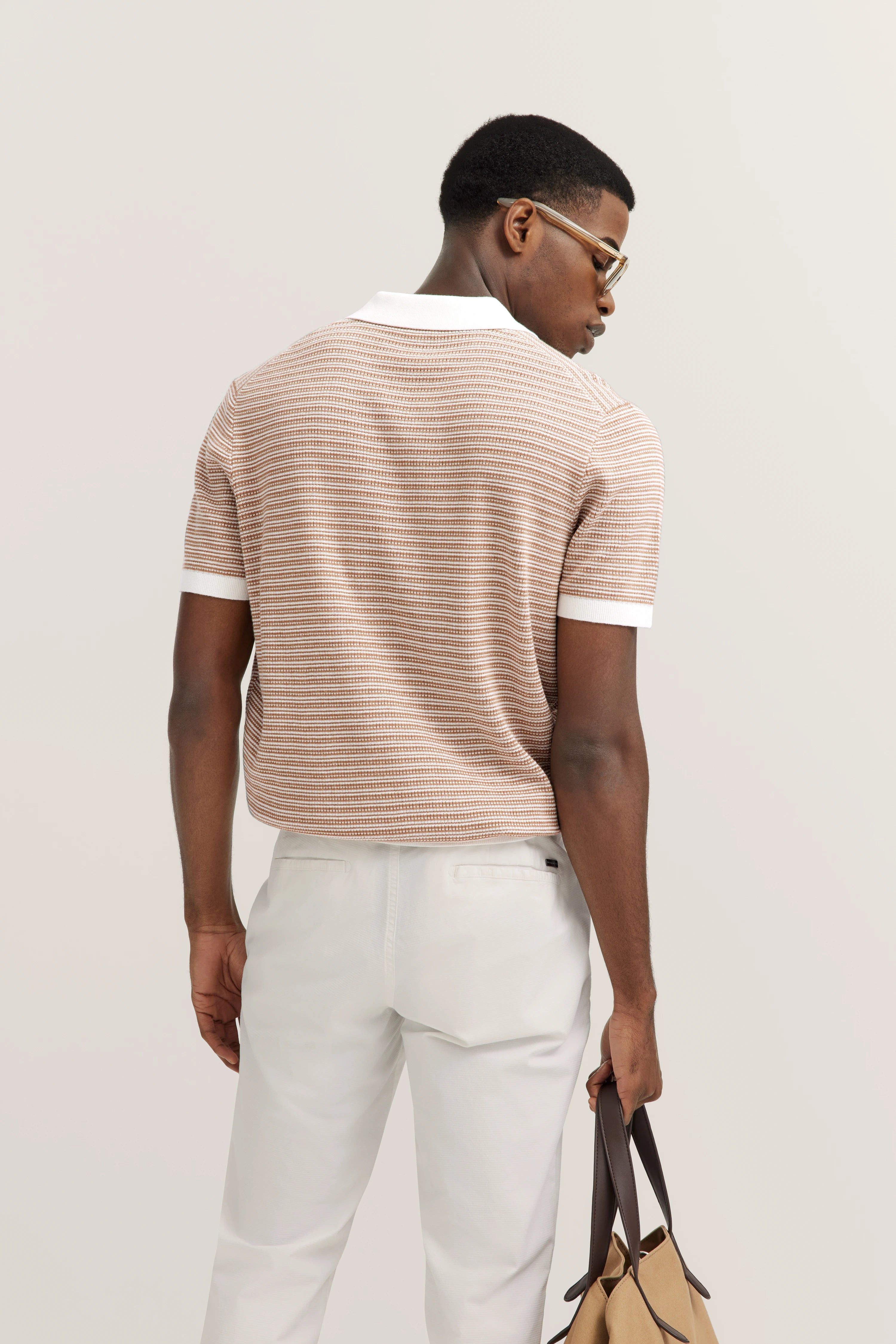 bugatti model in beige striped polo, white pants, back view, holding a bag.