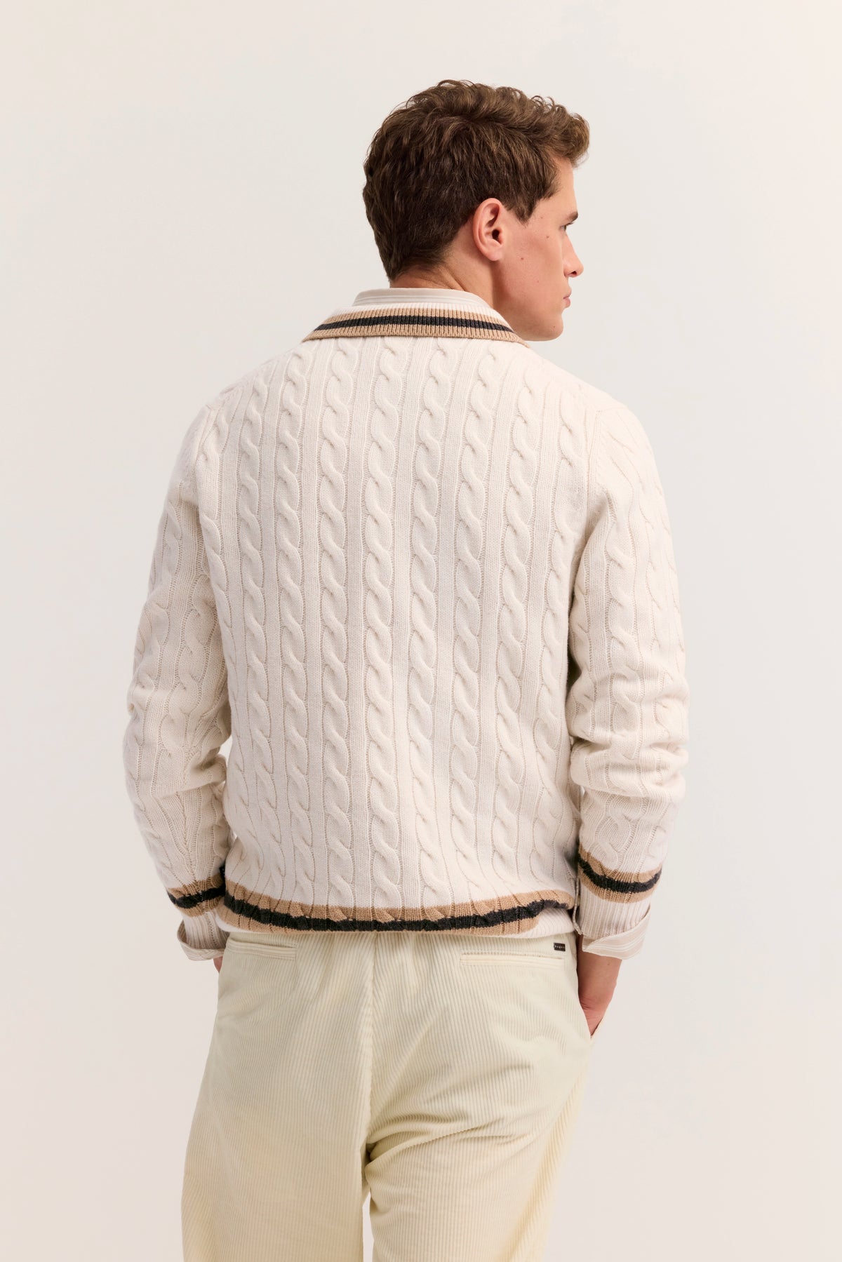 Model in cremefarbenem Strickpullover mit Zopfmuster und gestreiftem Kragen, Beige Hose, Rückansicht.