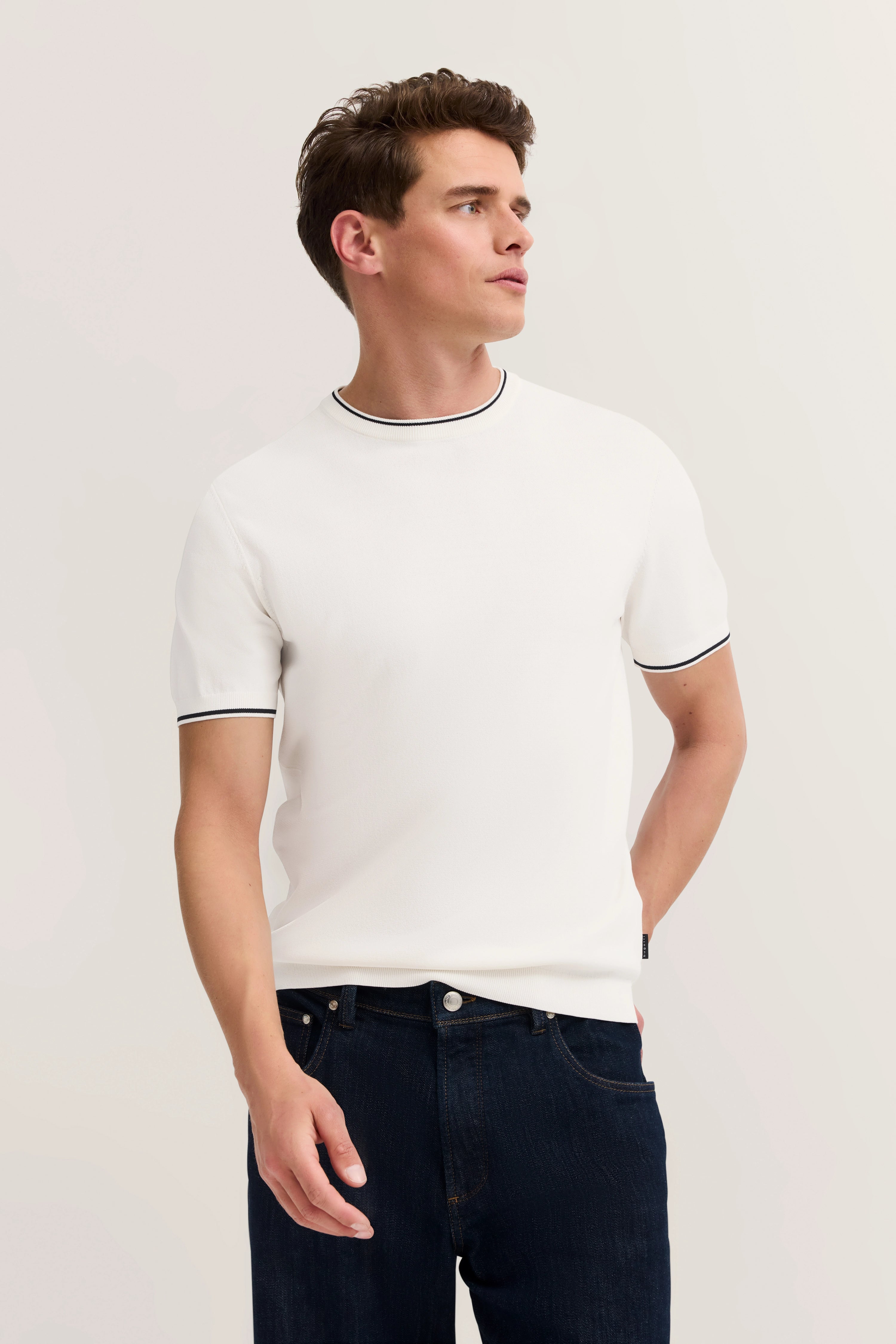 White bugatti T-shirt, black trim, dark blue jeans, model standing sideways in a relaxed pose.