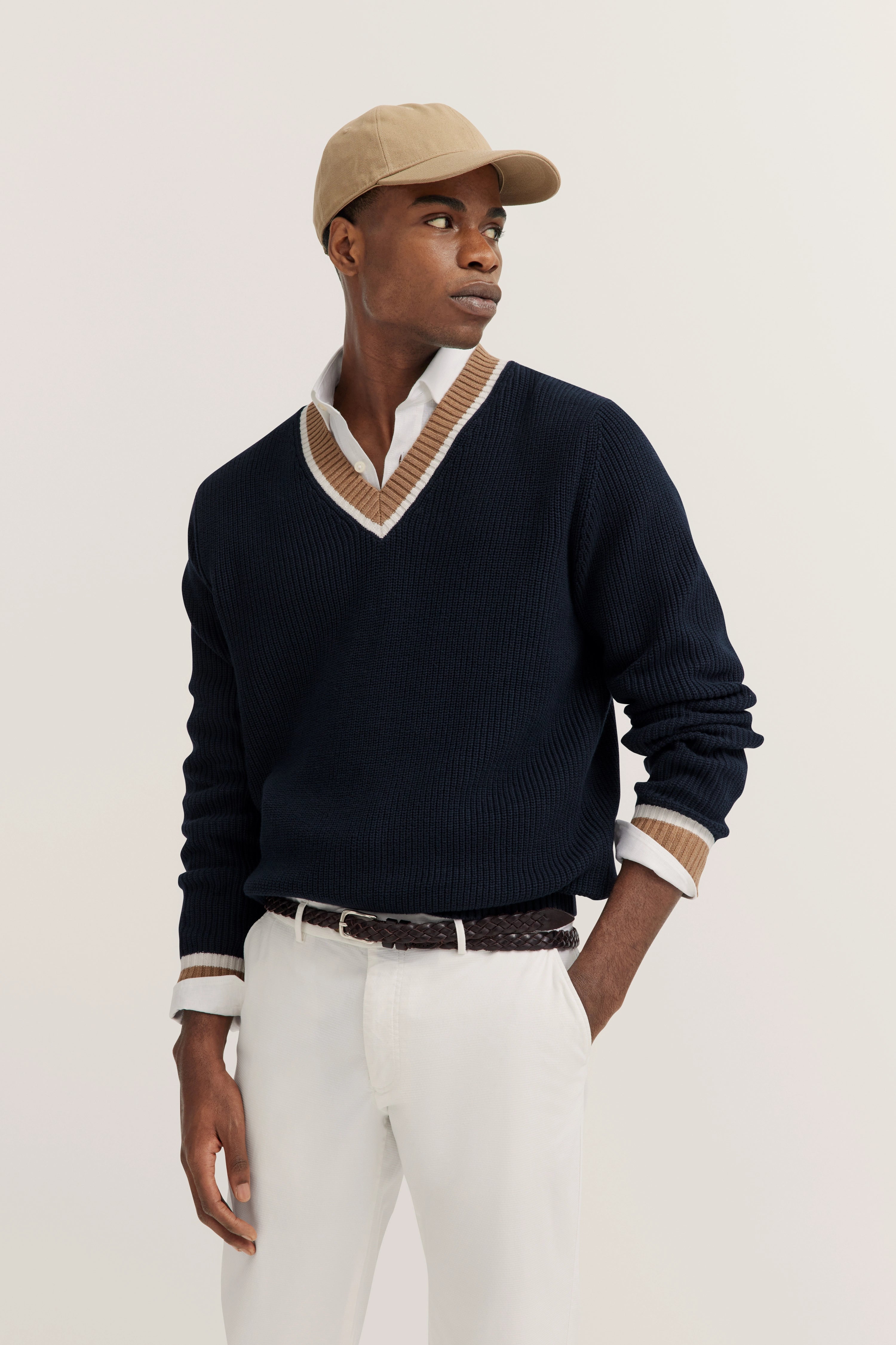 Model in beige pet, donkerblauwe trui met beige kraag en wit overhemd, poseert nonchalant.