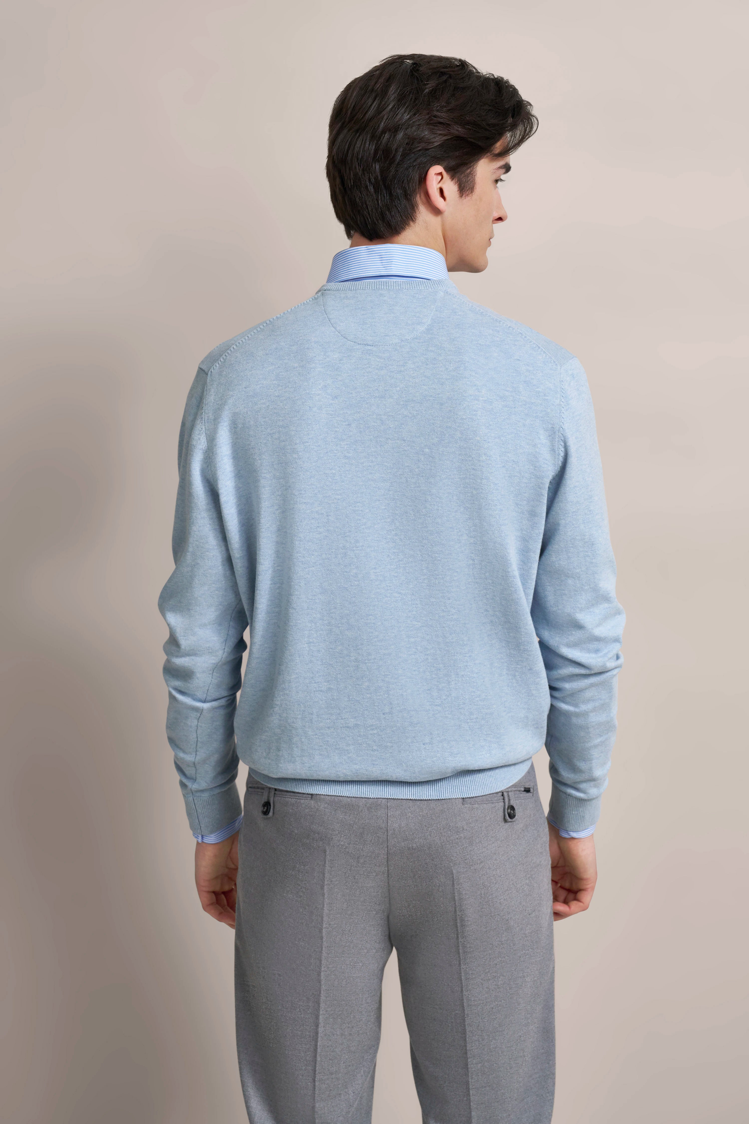 Rückansicht: Model in hellblauem Pullover, grauer Hose, steht entspannt. Posing: Rückenansicht.