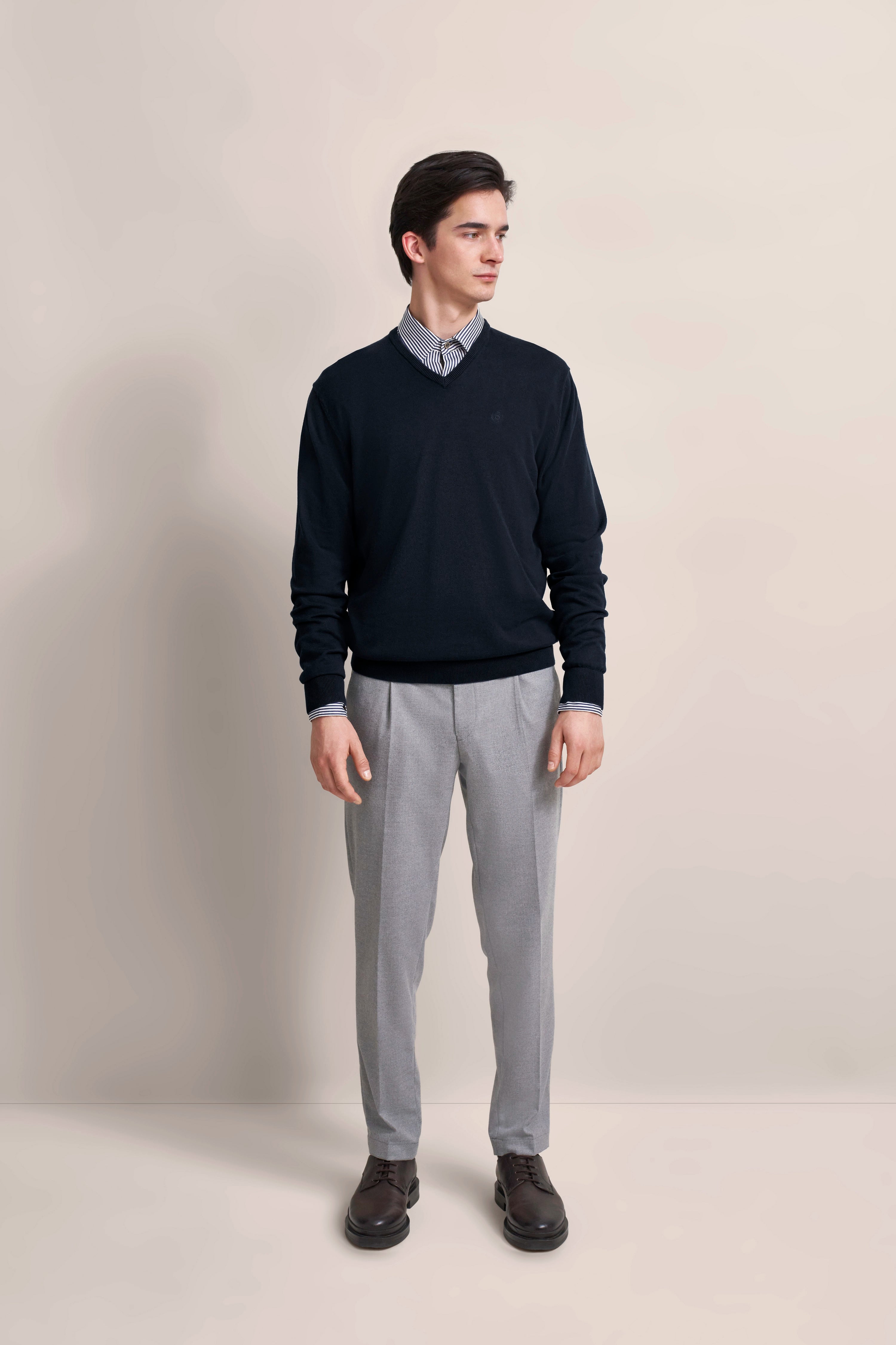 Model in schwarzem Pullover und grauer Hose, steht leger, trägt Hemd mit Streifen.
