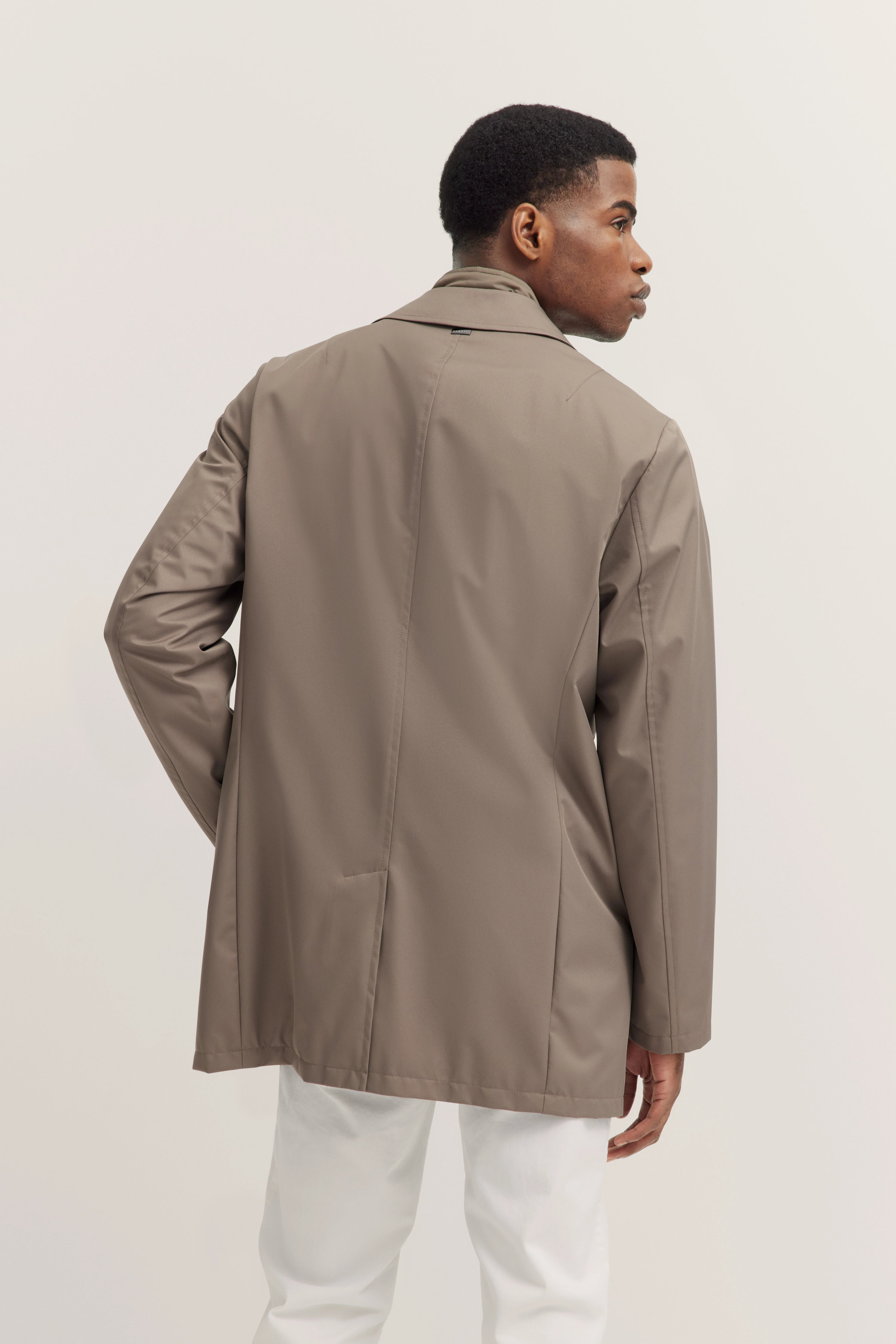 Model in taupefarbener bugatti-Jacke, Rückansicht, kombiniert mit weißen Hosen, entspannte Pose.