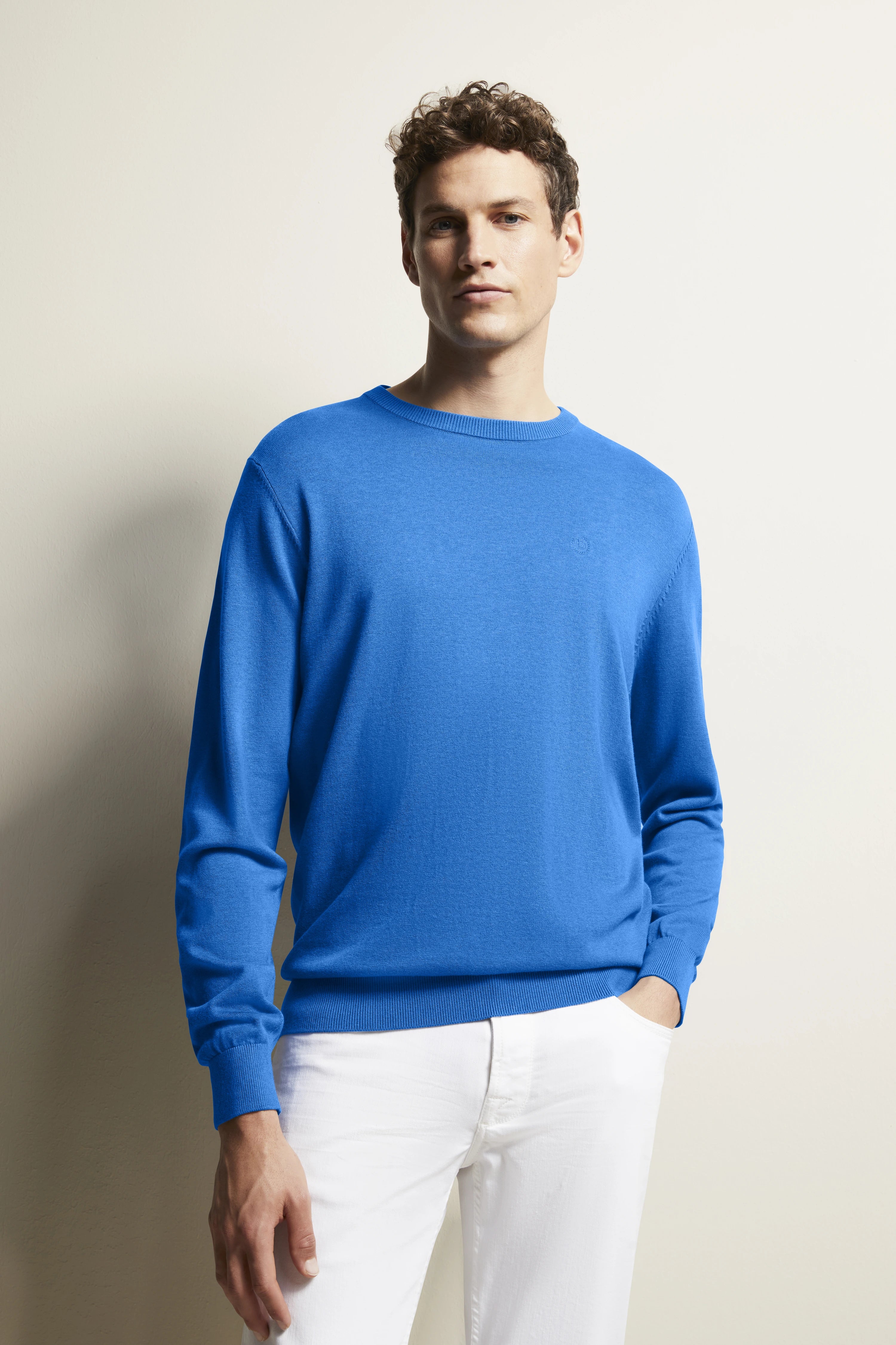 Model trägt blauen bugatti Pullover und weiße Hose, legeres Posing mit Hand in der Tasche.