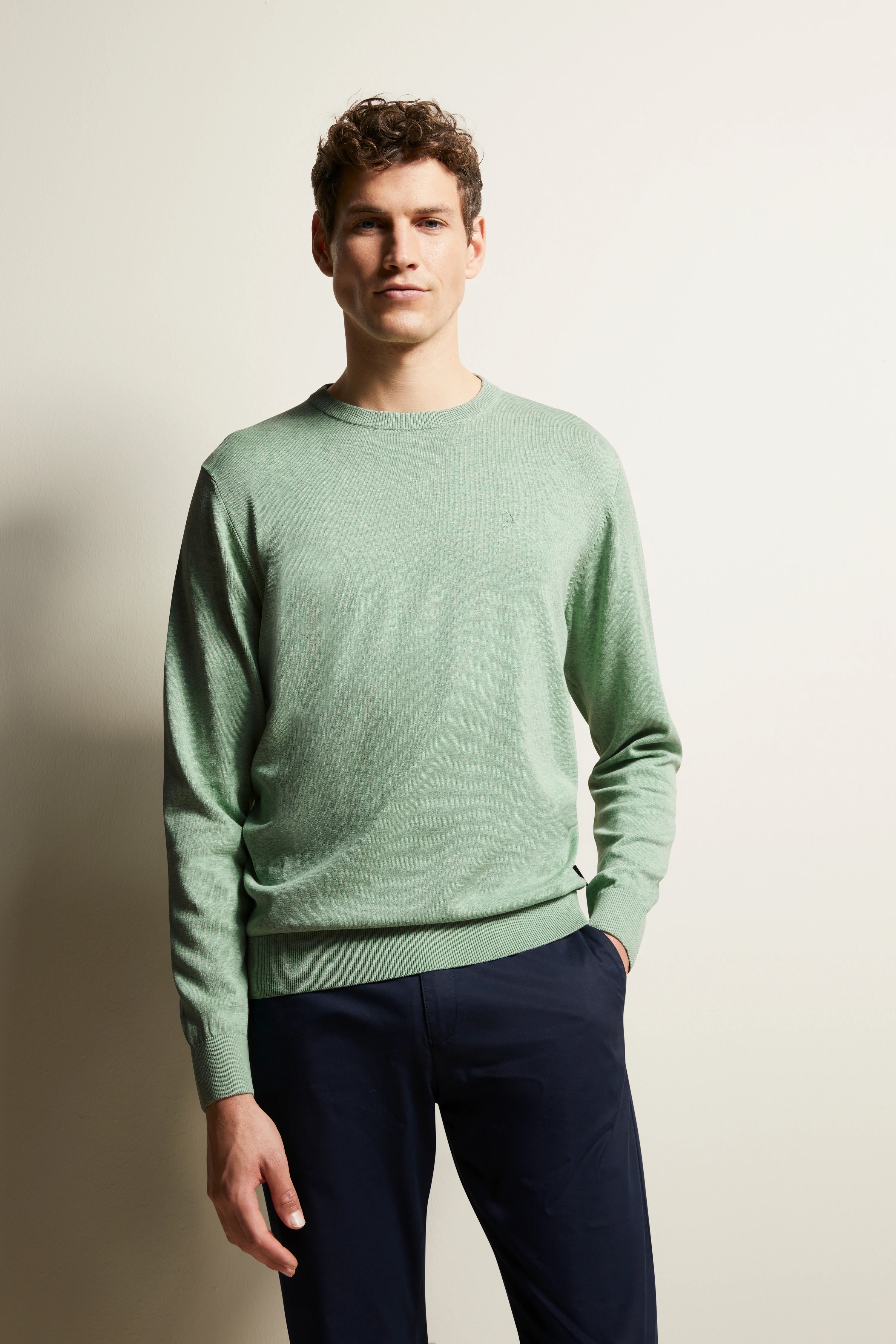 Bugatti Modelbild: Mann trägt mintgrünen Pullover, blaue Hose, lässige Pose, Hände in den Taschen.