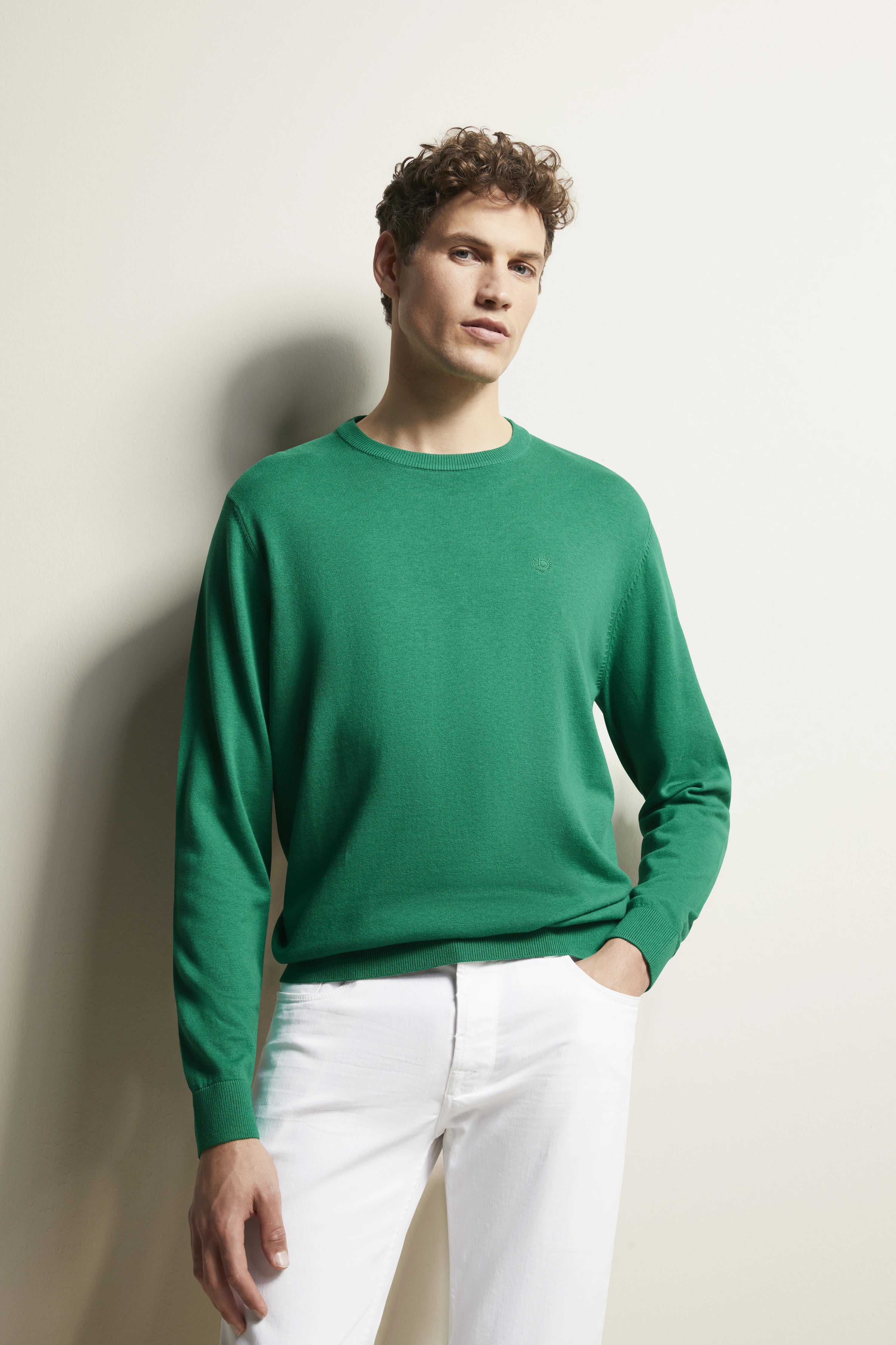 Model trägt grünen Pullover und weiße Hose von bugatti, locker stehend mit Hand in der Tasche.